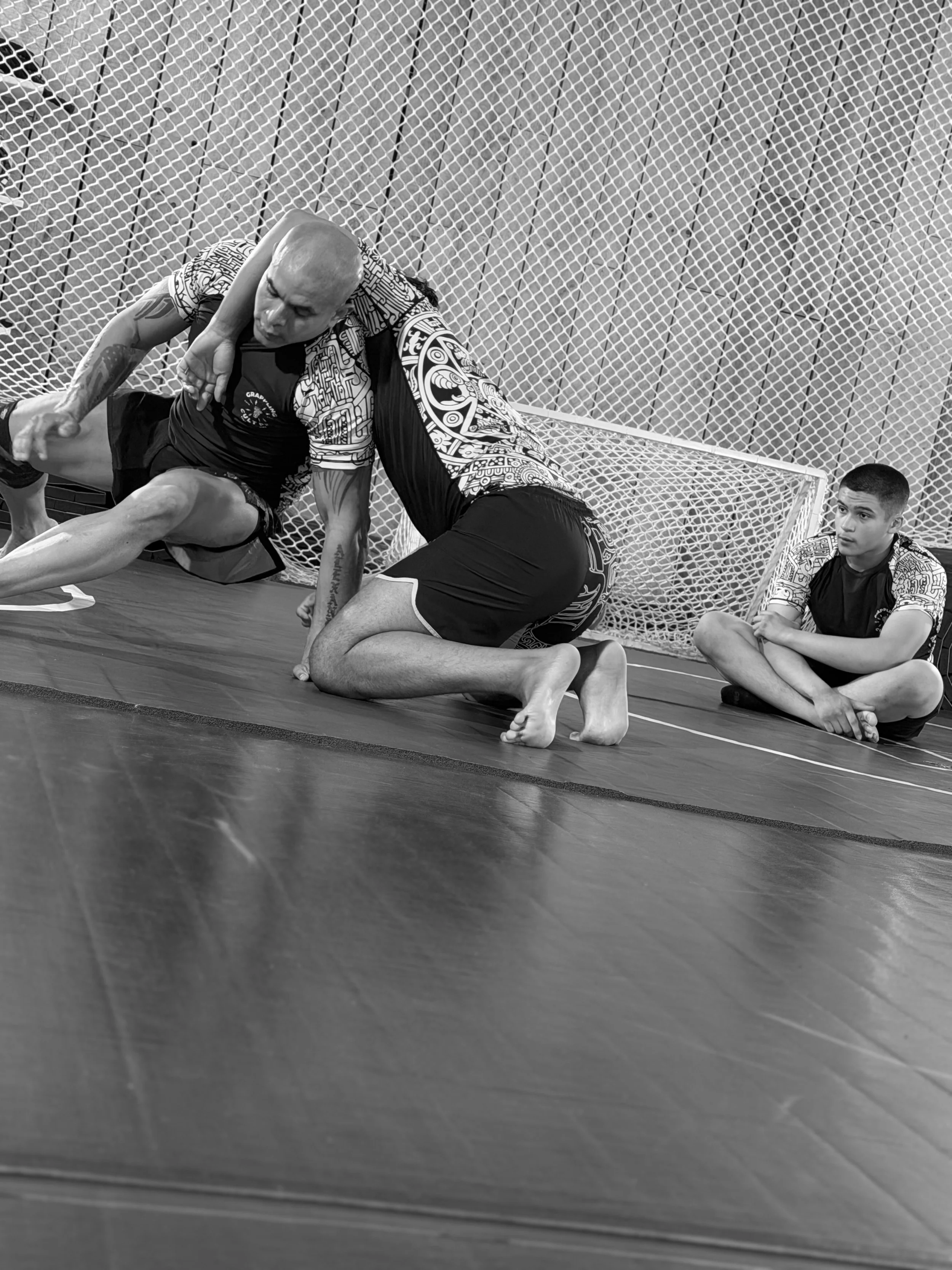 Two men practicing Brazilian Jiu-Jitsu on a mat, with a young boy sitting against the wall observing in the background.