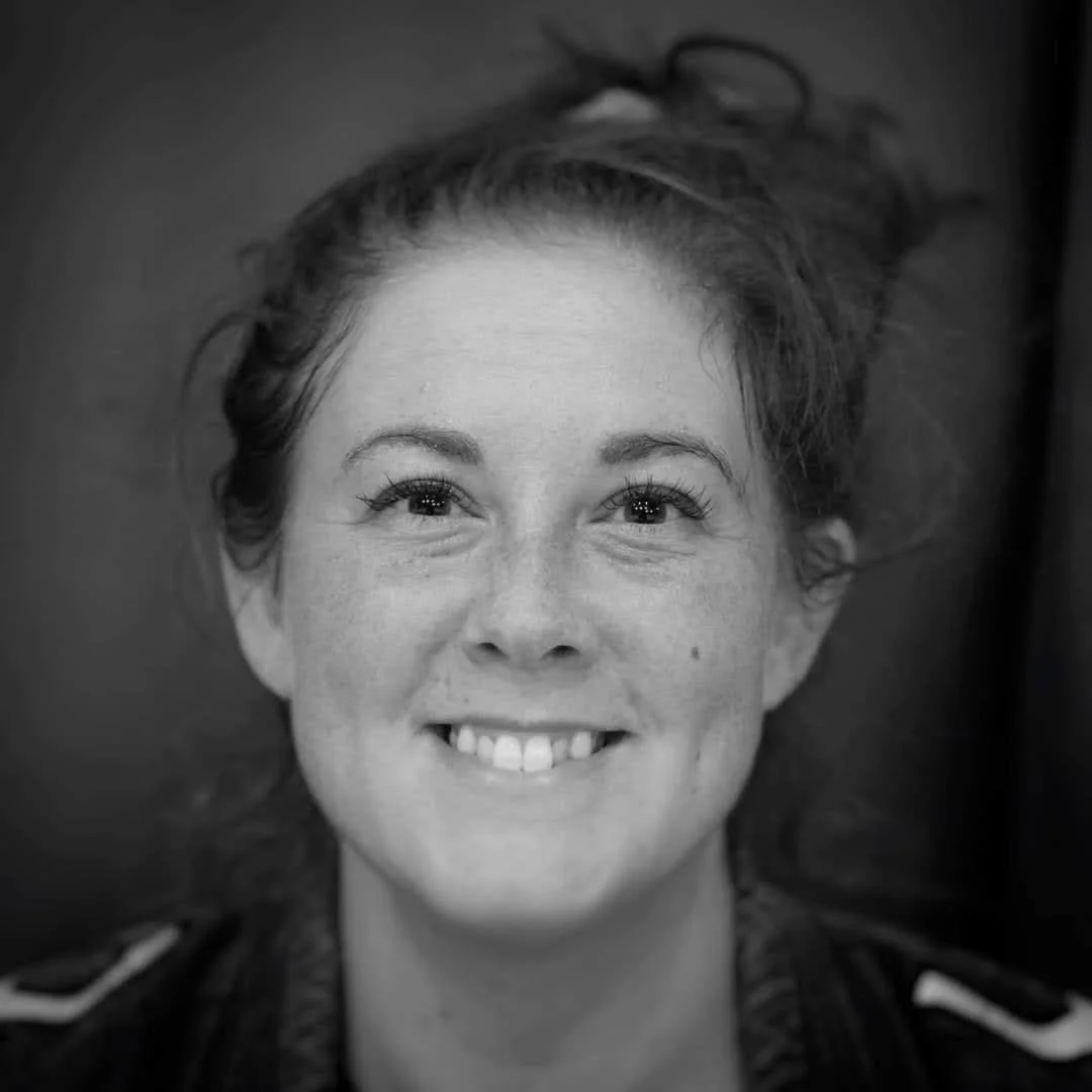 Black and white close-up portrait of a smiling woman with curly hair tied up in a bun, wearing a jacket with a visible collar.