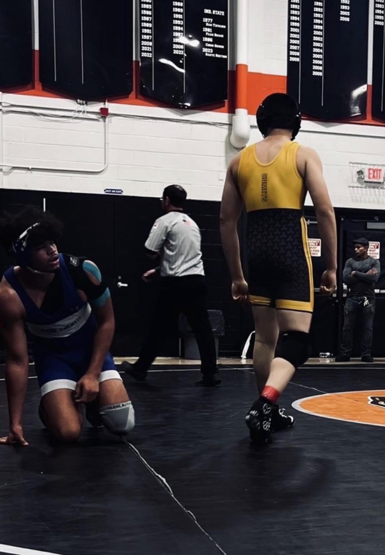 Wrestlers on the mat in a gym, with one kneeling and the other walking away, while referees and spectators are in the background, and scoreboard boards hanging on the wall.