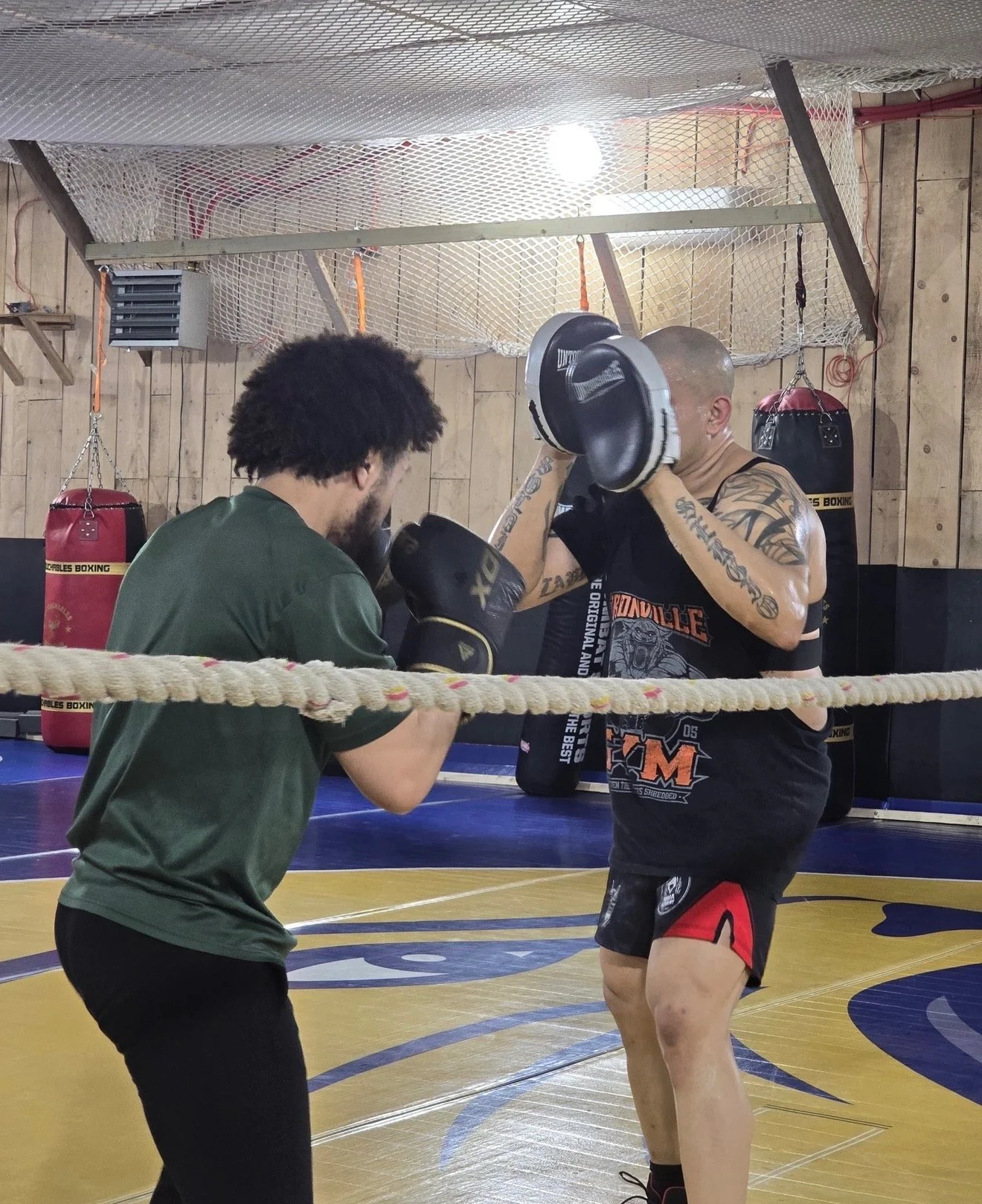 Two men are sparring in a boxing gym; the man on the left is practicing punches while the man on the right is holding focus mitts. The gym has a boxing ring, punching bags, and wood panel walls.