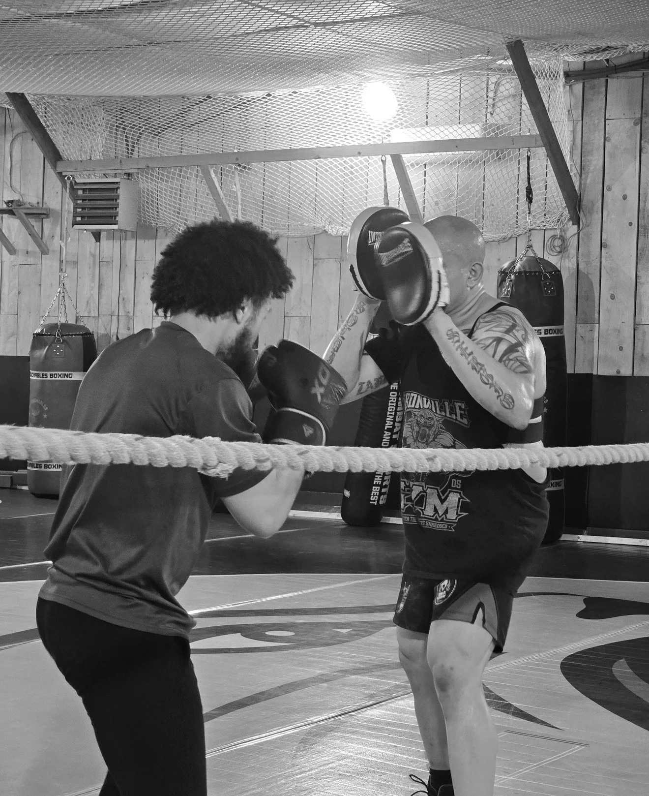 Two men practicing boxing in a gym. One man is delivering a punch while the other is blocking with gloves. The gym has wooden walls, hanging punching bags, and a netted ceiling.
