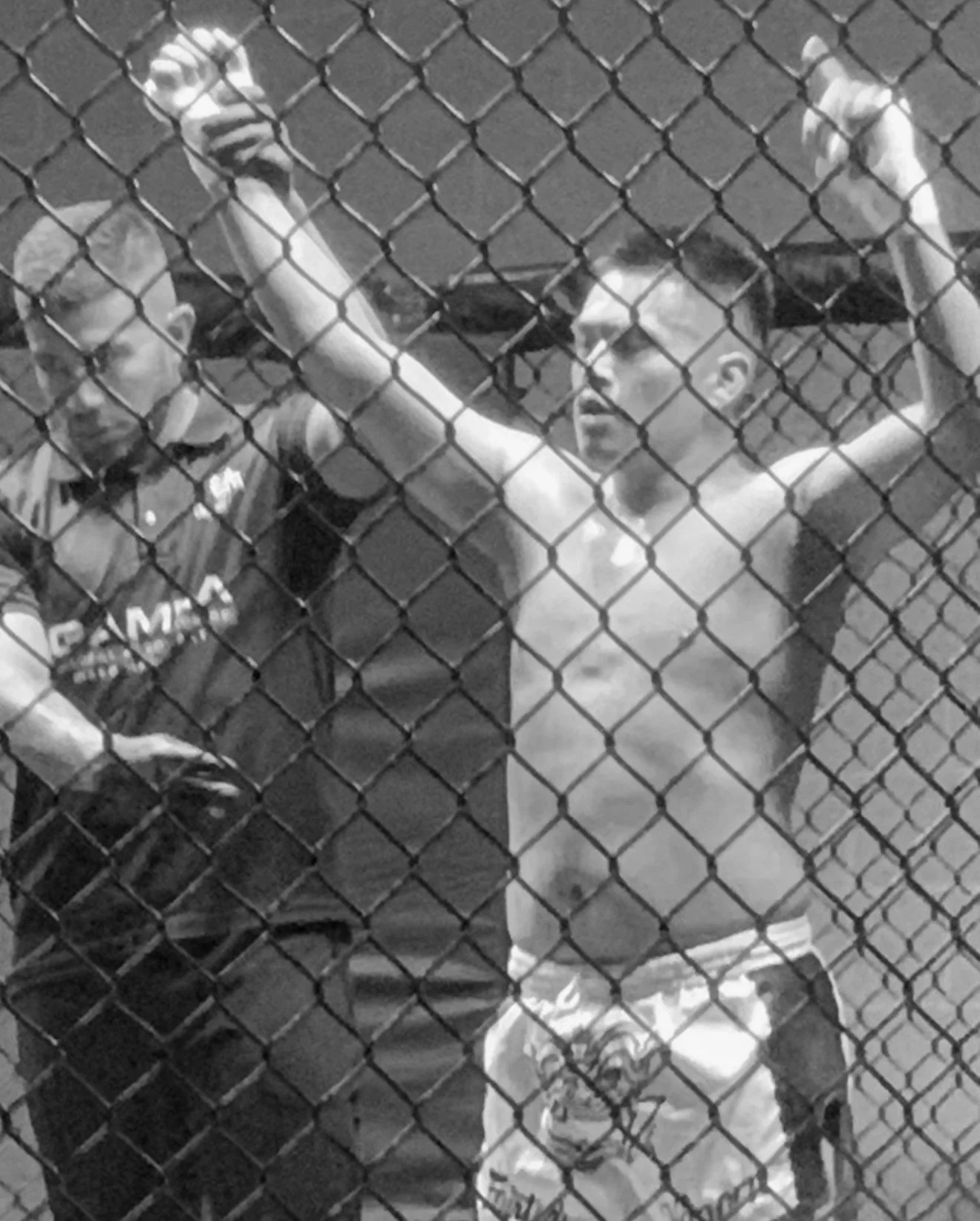 Two young boys behind a chain-link fence, one perspiring, with the other boy wearing glasses and a sports shirt. The boy on the right has hands resting on the fence.