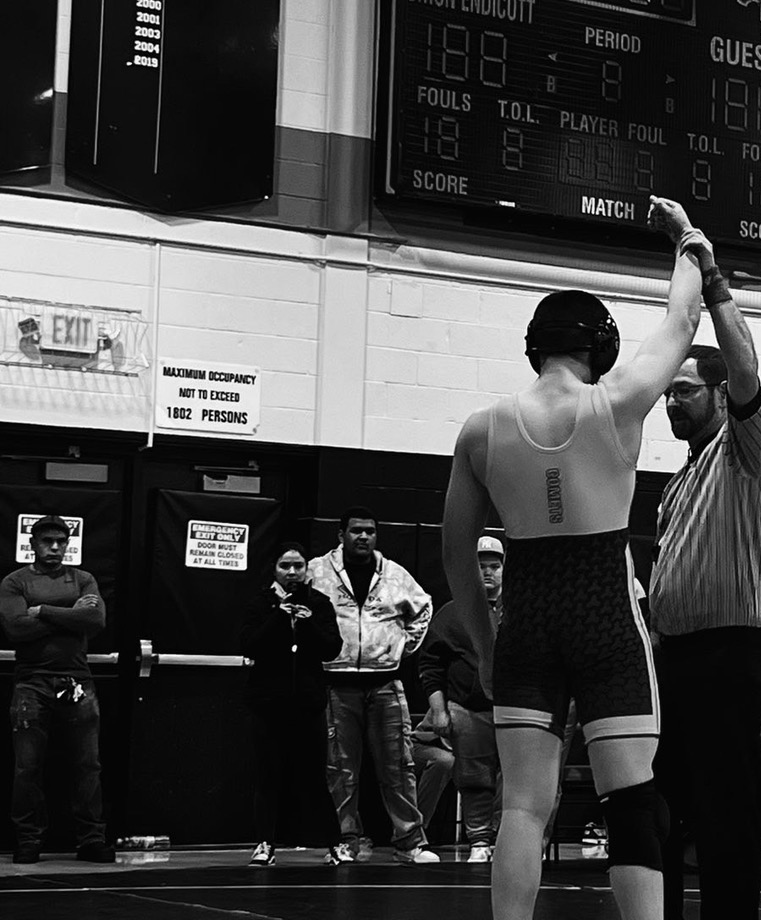 A wrestler in black and gray attire with a tattoo on the back receives a handshake from a referee during a match in a gymnasium. Several spectators watch from the background, and a scoreboard is visible above showing game details.