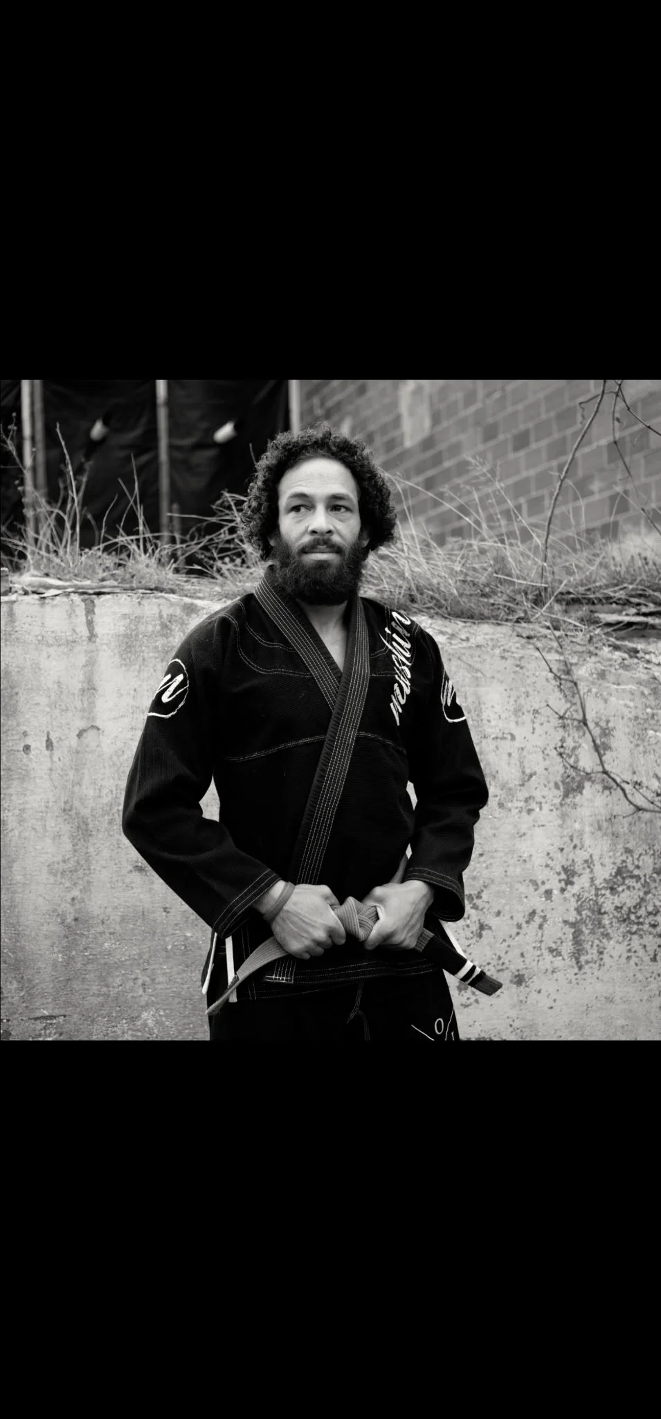 A man in a Brazilian Jiu-Jitsu gi with a black belt stands outdoors against a concrete wall, holding his belt with both hands, with a brick wall and some dried plants in the background.