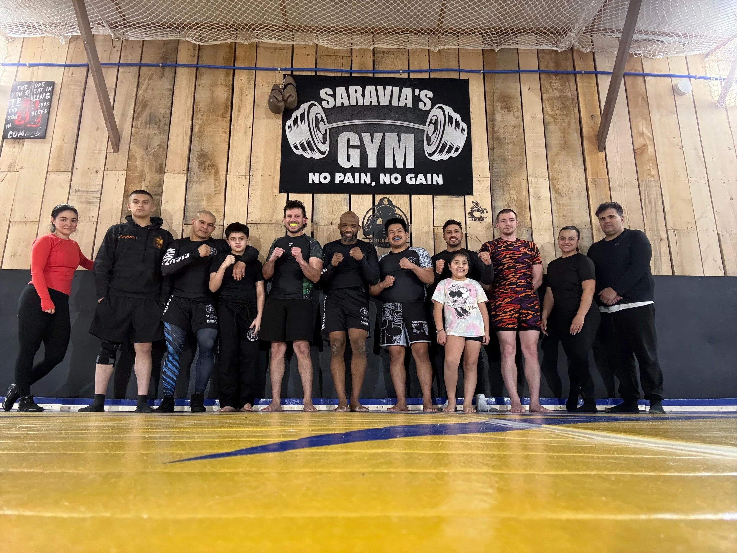 Group of people in athletic clothing standing on a gym floor in front of a wooden wall with a banner that says "Saravia's Gym No Pain No Gain" and a large barbell graphic. Some are smiling and making fists, indicating a fitness or martial arts class.