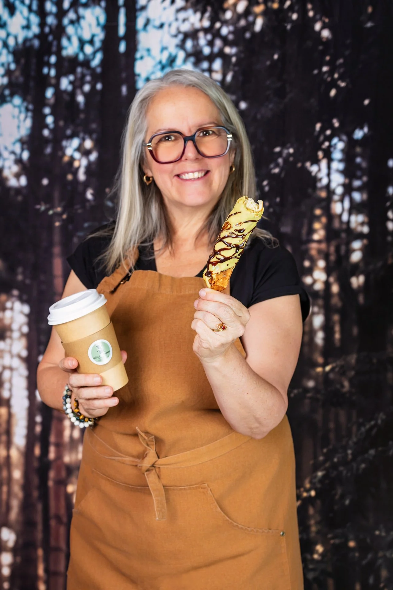 Une femme souriante avec des lunettes, portant un tablier en cuir marron, tient une tasse à café en carton dans une main et une grande crème glacée en cornet avec du chocolat fondu dans l'autre. Elle se tient devant un fond d'arbres dans la forêt.