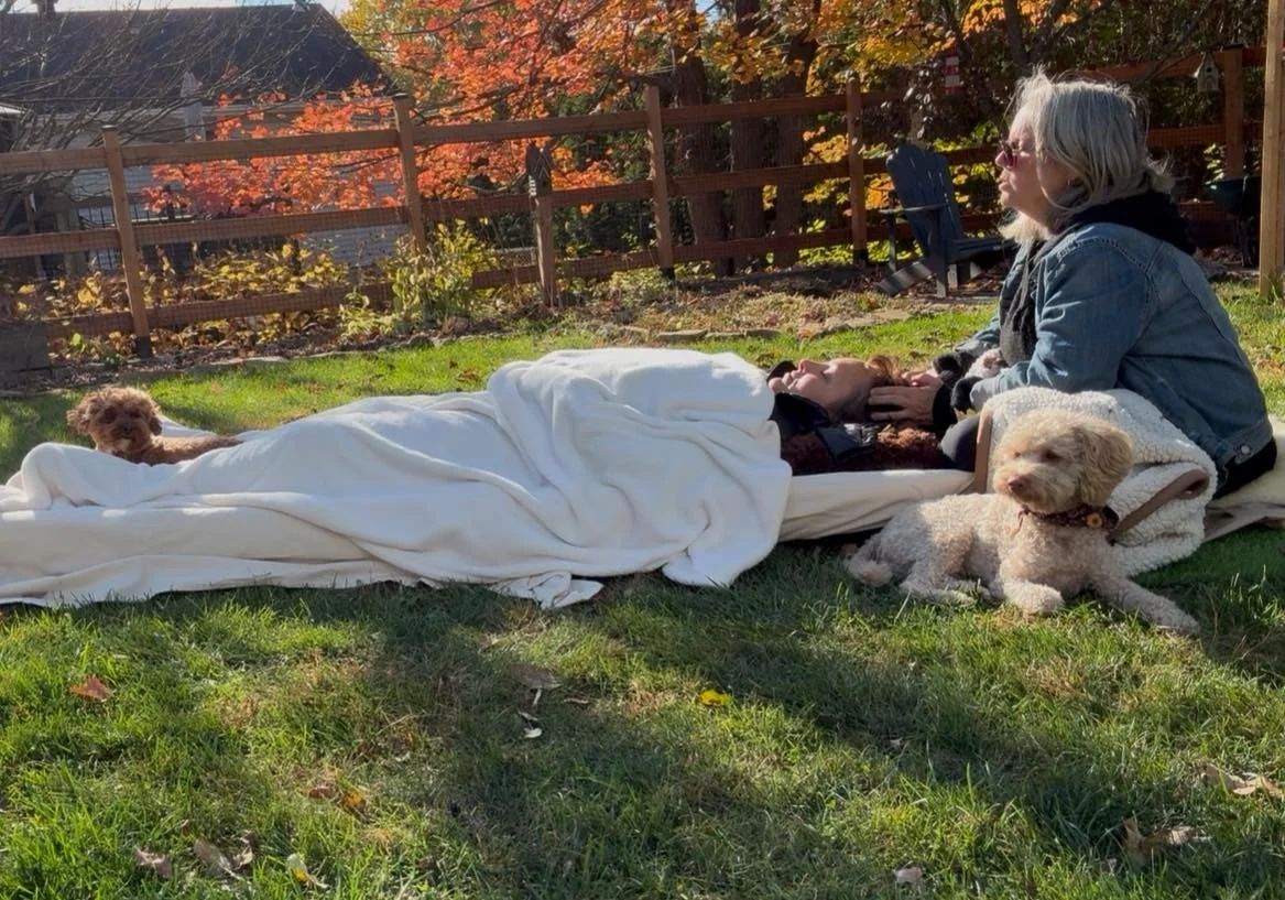 Une femme allongée sur l'herbe avec deux chiens, une autre femme assise à côté avec un chien, dans un jardin en automne.
