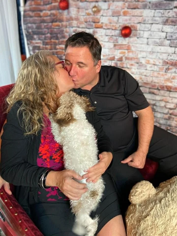 Un homme et une femme s'embrassent sur la bouche dans un salon, avec deux chiens à proximité, en arrière-plan un mur en brique décoré de boules de Noël rouges.