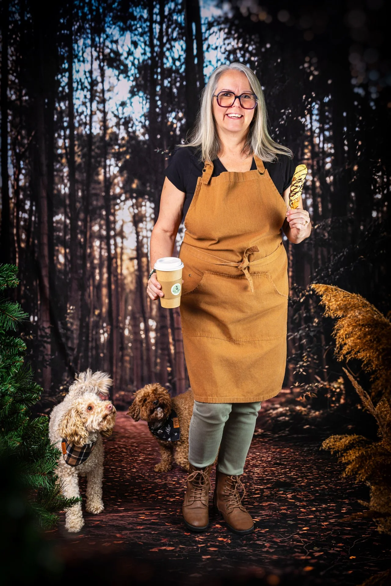 Femme âgée tenant un café à emporter et une pâtisserie, accompagnée de deux chiens, dans un décor de forêt