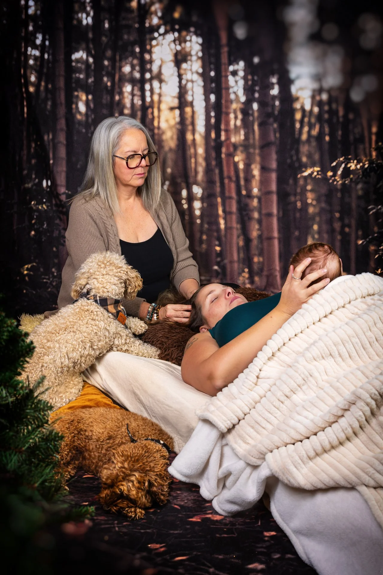 Une femme allongée sur un lit, recevant un massage de la part d'une autre femme dans une forêt. Deux chiens sont présents : un canine puli et un autre petit toutou, posés sur le lit et le sol, dans un environnement naturel.