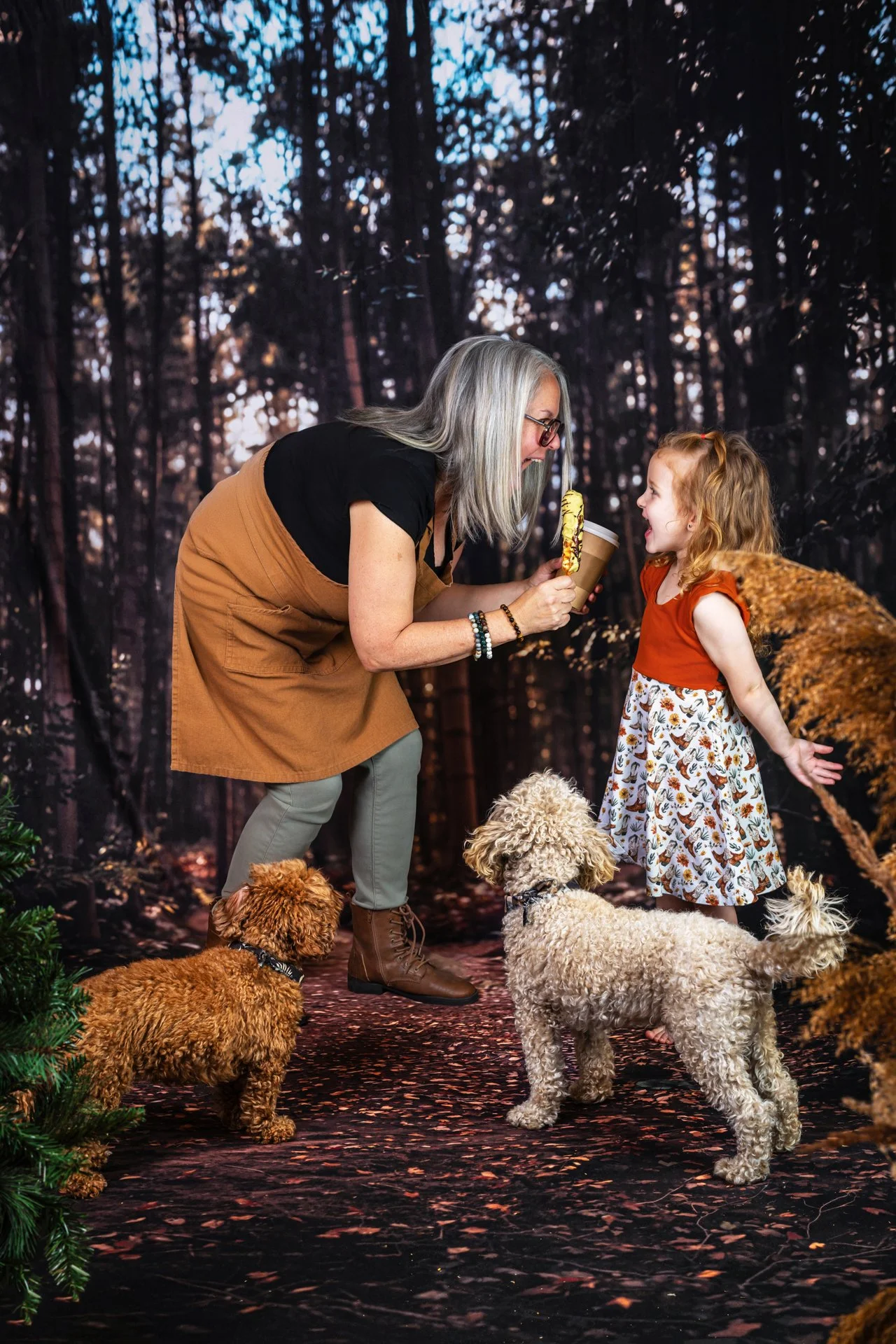 Une femme avec des cheveux gris, portant un tablier brun, une robe noire et des bottes marron, offre une glace à une petite fille rousse dans une forêt lointaine. Deux chiens, un petit caniche brun et un autre crème, se tiennent près d'eux, tandis que la petite fille sourit. L'arrière-plan est une forêt dense avec des arbres.