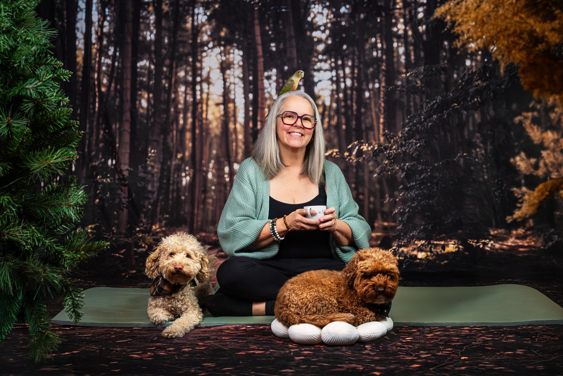 Une femme avec des cheveux gris, portant des lunettes, assise sur un tapis dans un décor de forêt, tenant une tasse. Elle est accompagnée de deux chiens, un à sa gauche et un autre devant elle, et a un perroquet posé sur sa tête. Le fond représente une forêt avec des arbres.