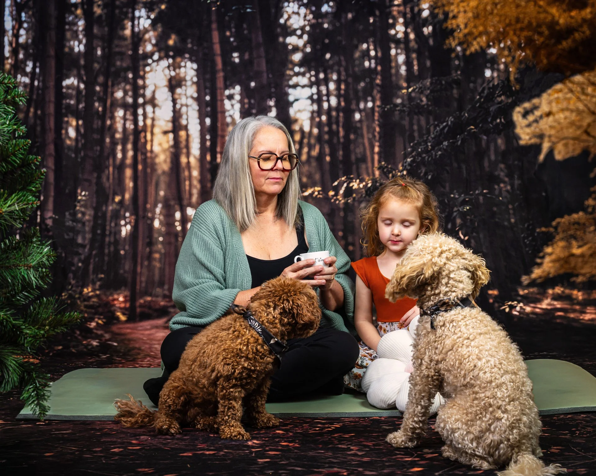 Une femme assise sur un tapis vert dans une forêt avec deux chiens et une jeune fille. La femme tient une tasse dans ses mains. La forêt en arrière-plan est sombre avec des arbres et une lumière douce.