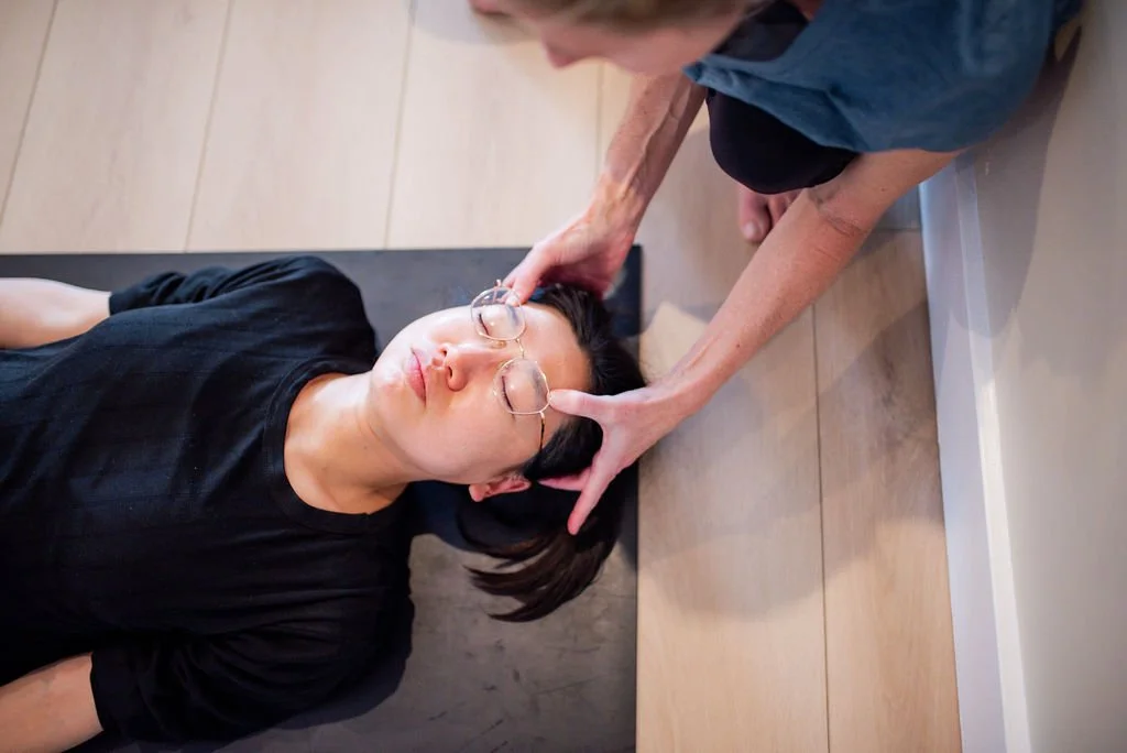A woman lying on the floor with her eyes closed, receiving a head massage from an older woman.