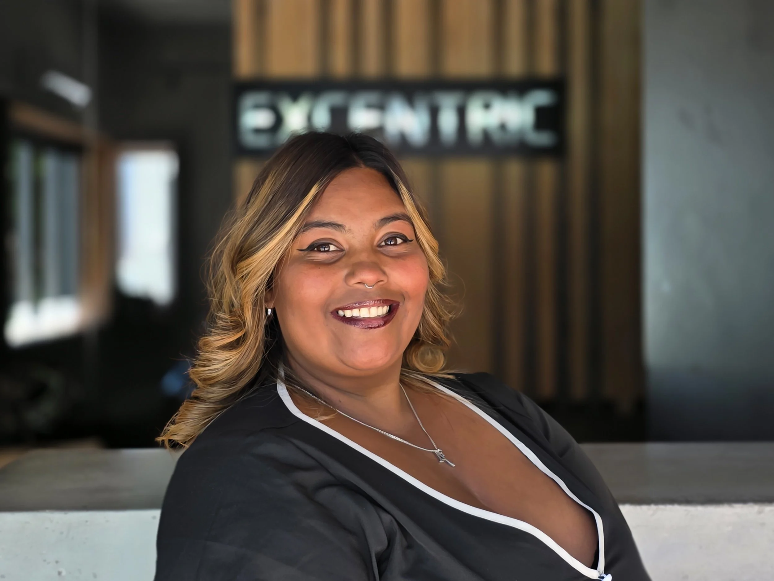 Smiling woman with shoulder-length wavy hair and a septum piercing, wearing a black top with white trim, in a modern interior space with a blurred background and a sign that reads 'EXPERIENCE' in the background.
