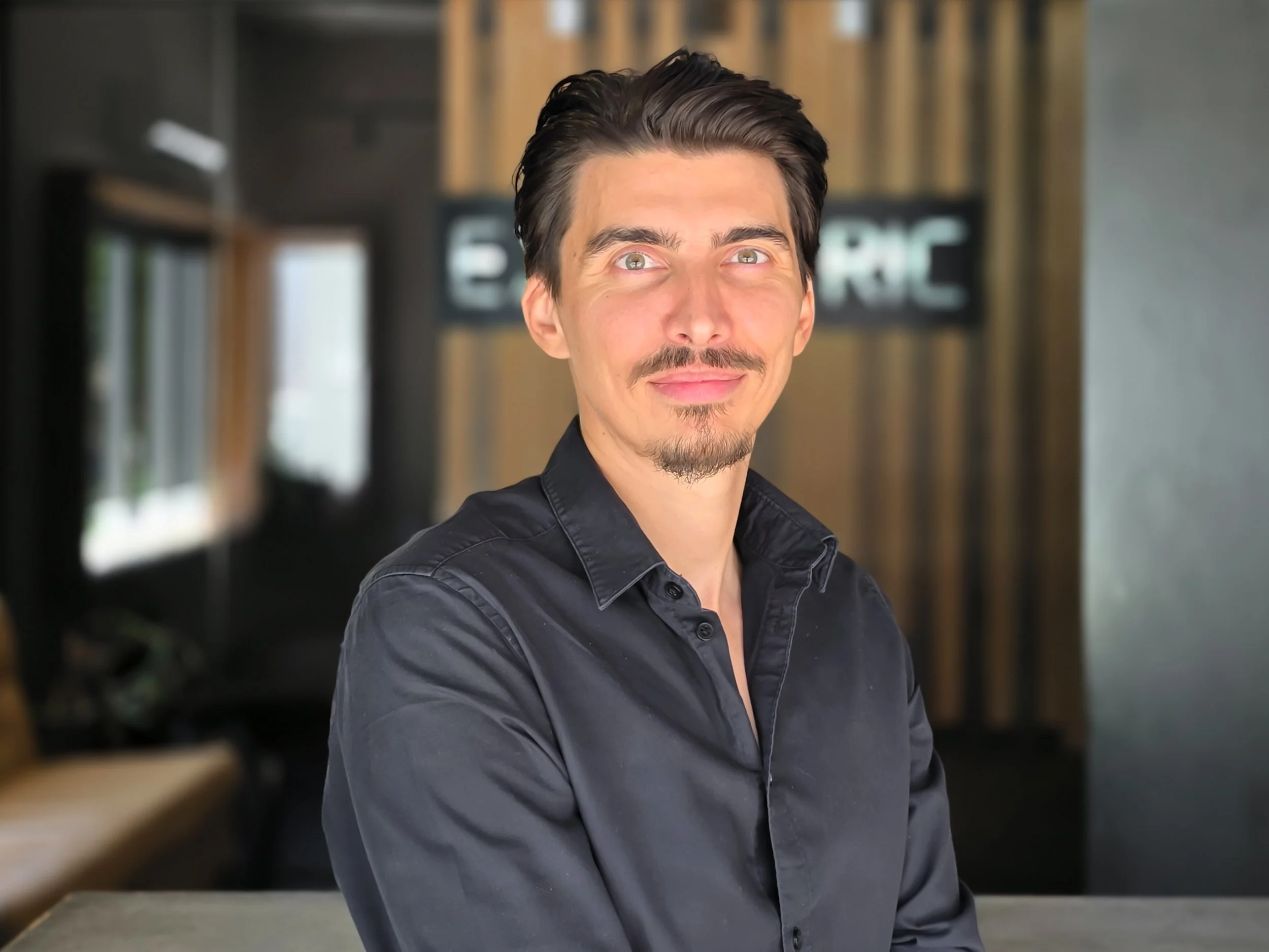 A man with dark hair, mustache, and goatee smiling and looking at the camera, wearing a black button-up shirt, in a modern indoor setting with wood accents and blurred background.