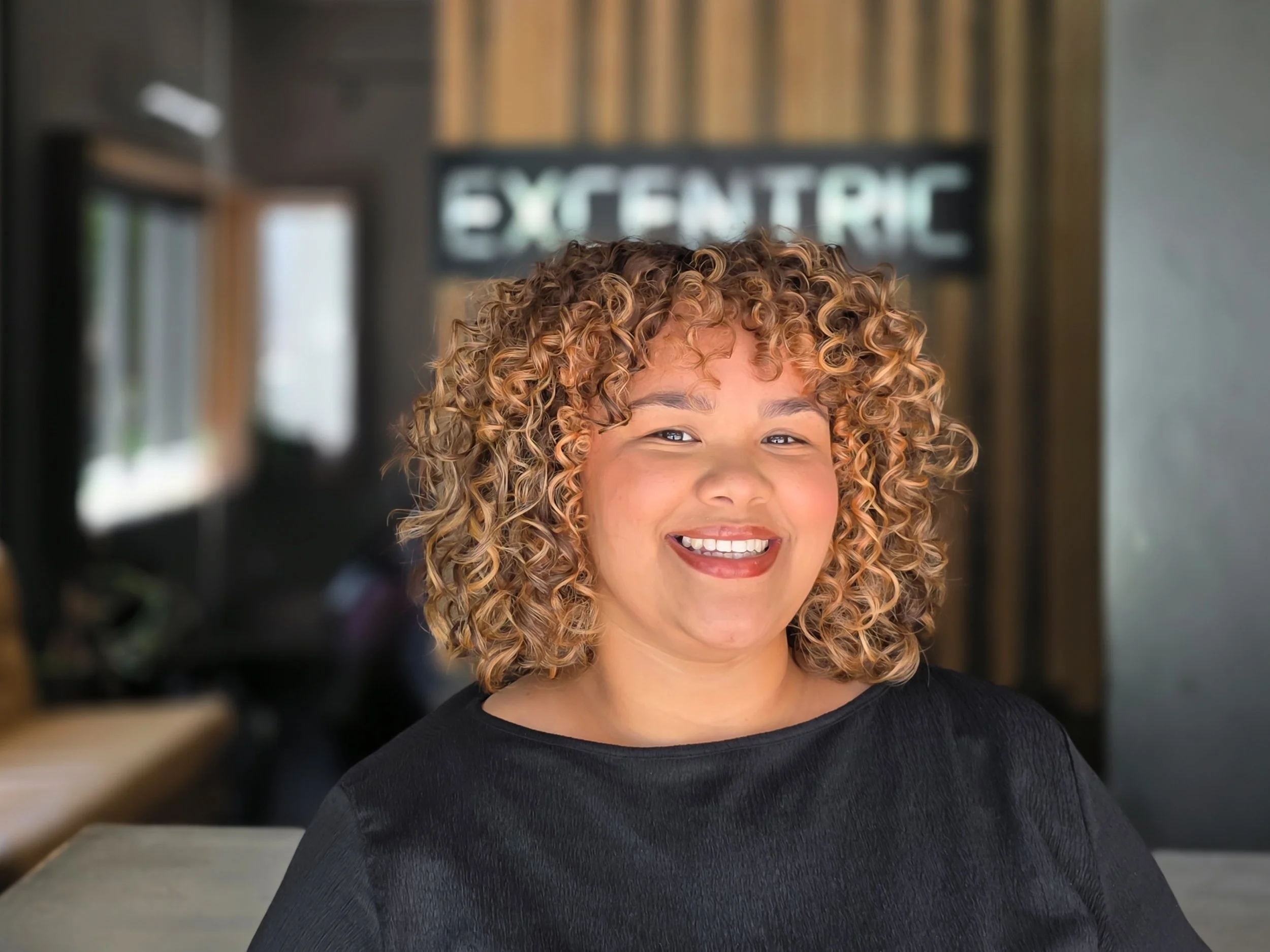 Close-up of a woman with curly, blonde hair smiling in a modern indoor space with a blurred background and a sign reading 'ELECTRIC'.