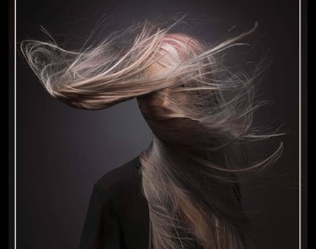 Close-up of a woman with long, flowing gray hair blowing across her face against a dark background.