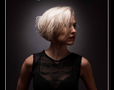 A woman with short platinum blonde hair wearing a black sleeveless top in a dark studio setting.