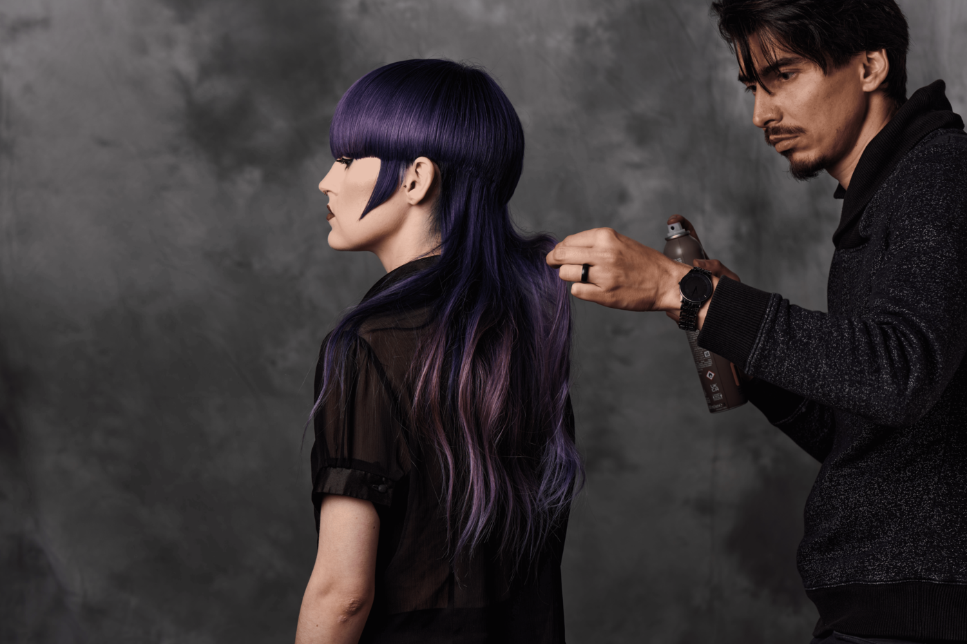 A hairstylist sprays hair of a woman with vibrant purple and blue hair, against a gray textured background.