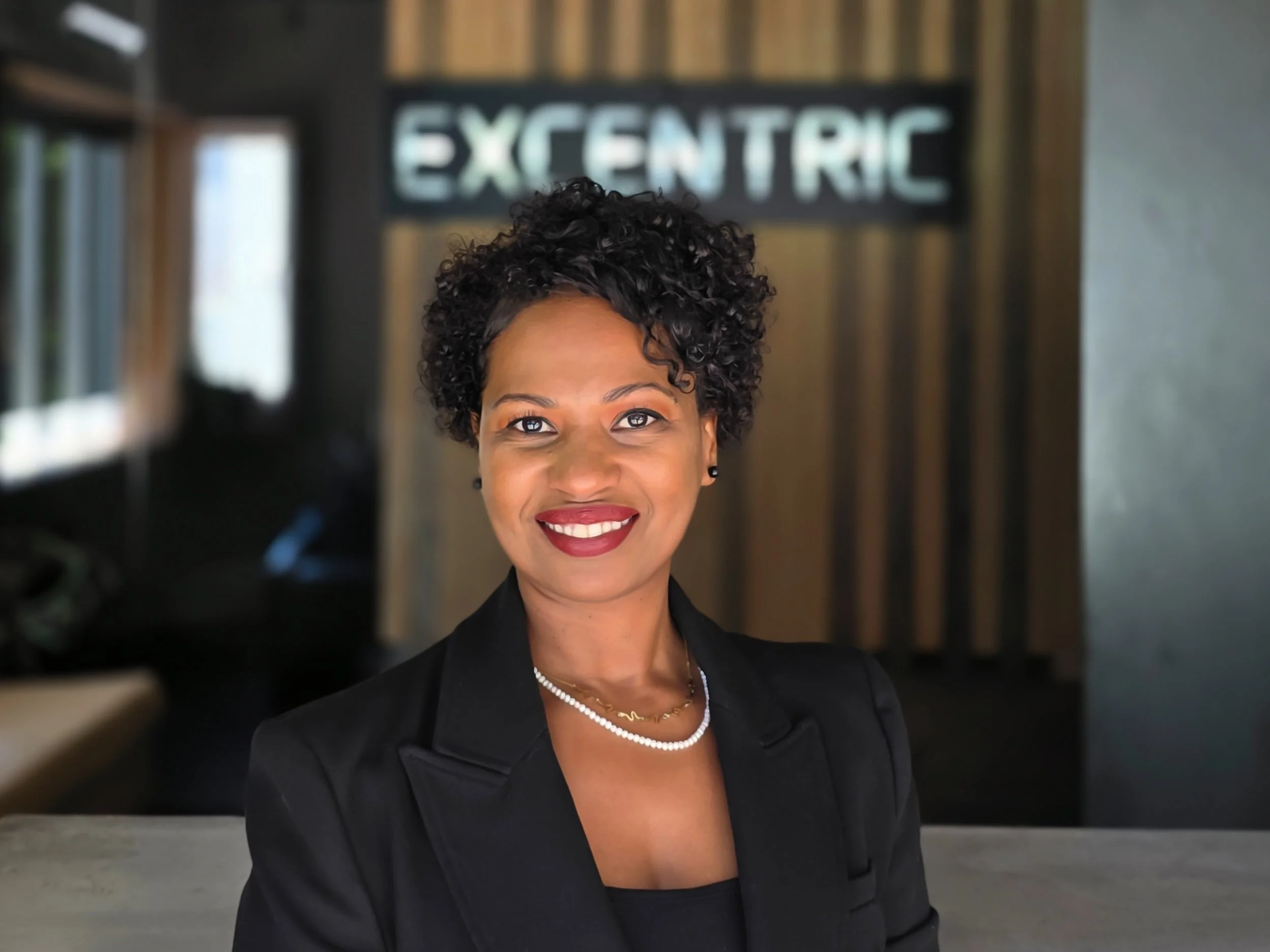 A woman smiling in a professional setting with a sign that says 'Excentric' in the background.