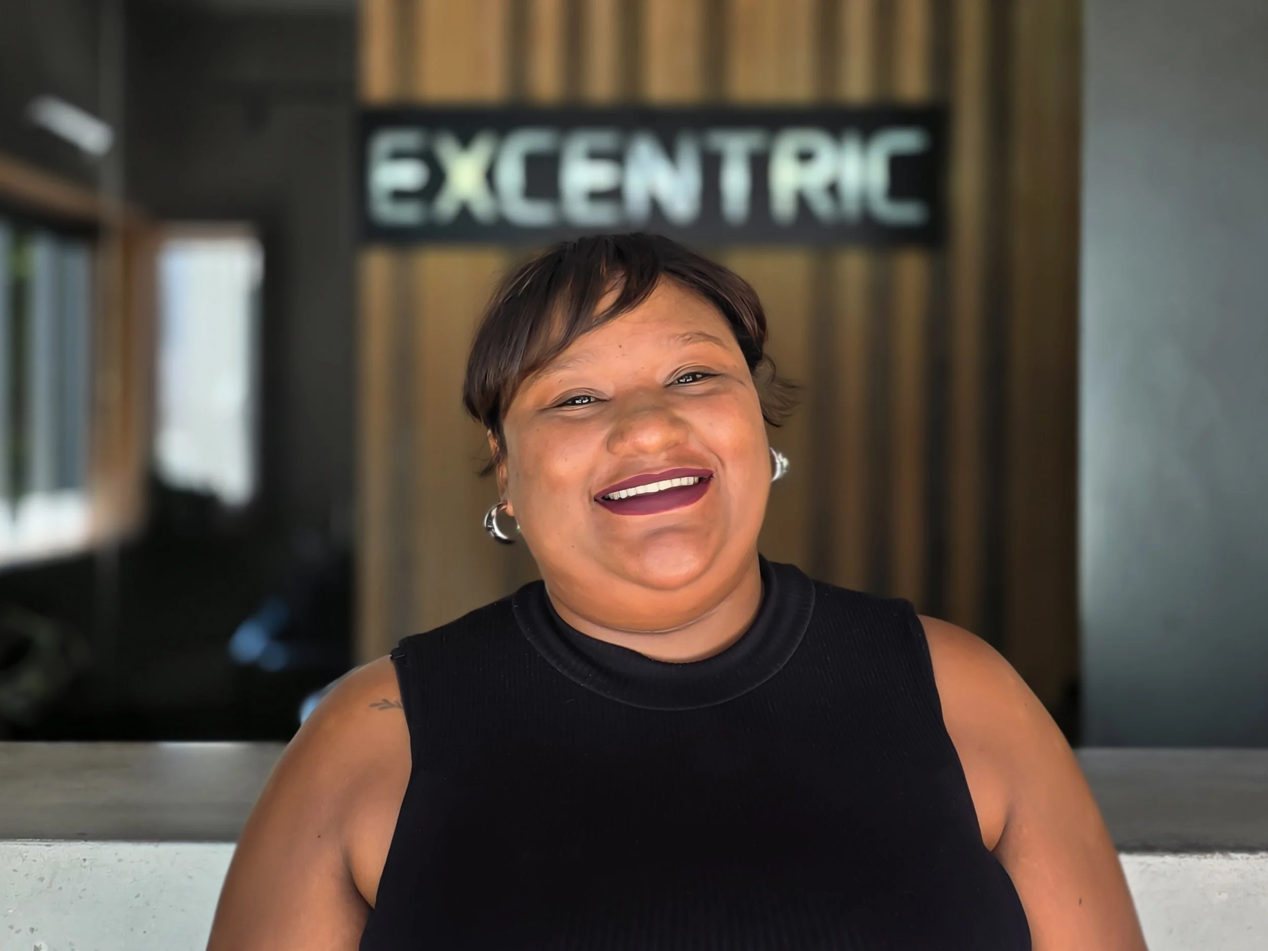 Portrait of a smiling woman with short hair, wearing a black sleeveless top, standing indoors with a blurred sign that reads 'EXCENTRIC' in the background.