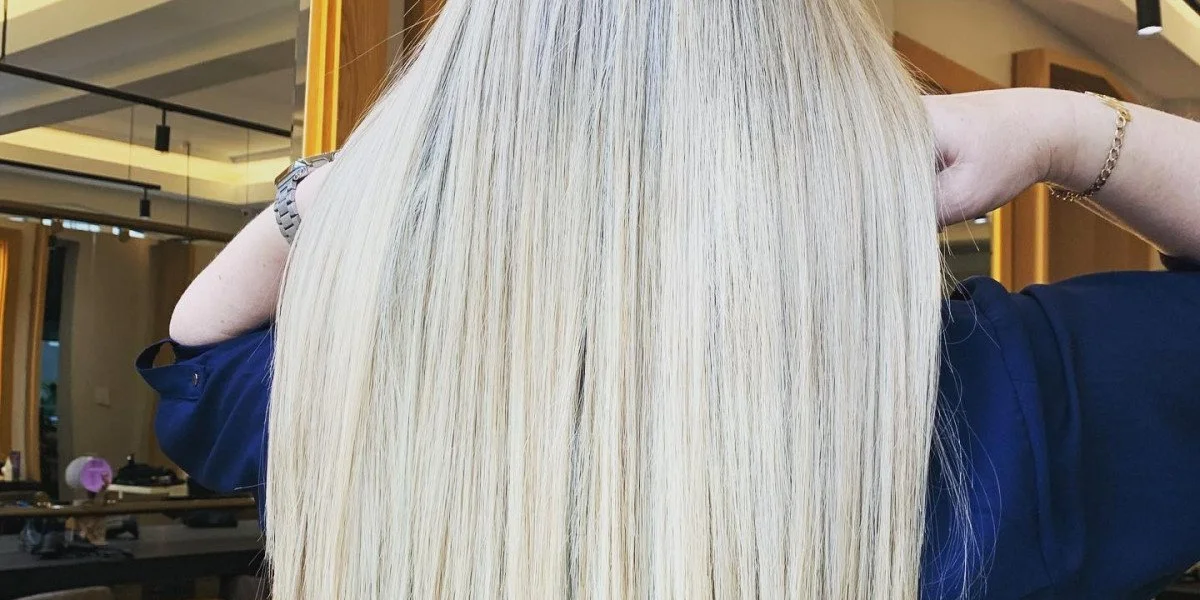 Person with long, straight, blonde hair, wearing a navy-blue shirt, in a salon setting.