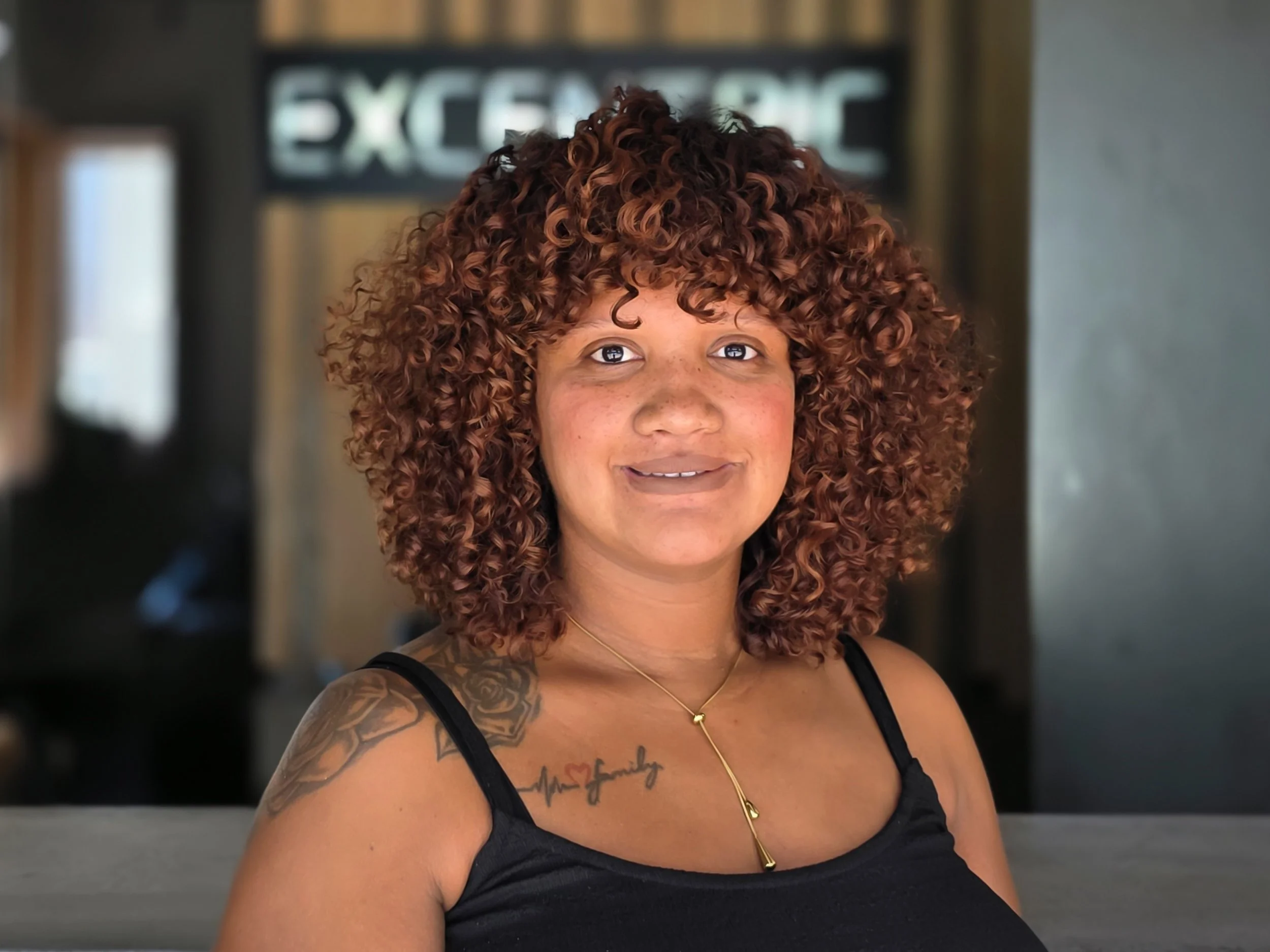 A woman with curly brown hair, wearing a black top, a gold necklace, and a tattoo on her shoulder. She is smiling and standing indoors.