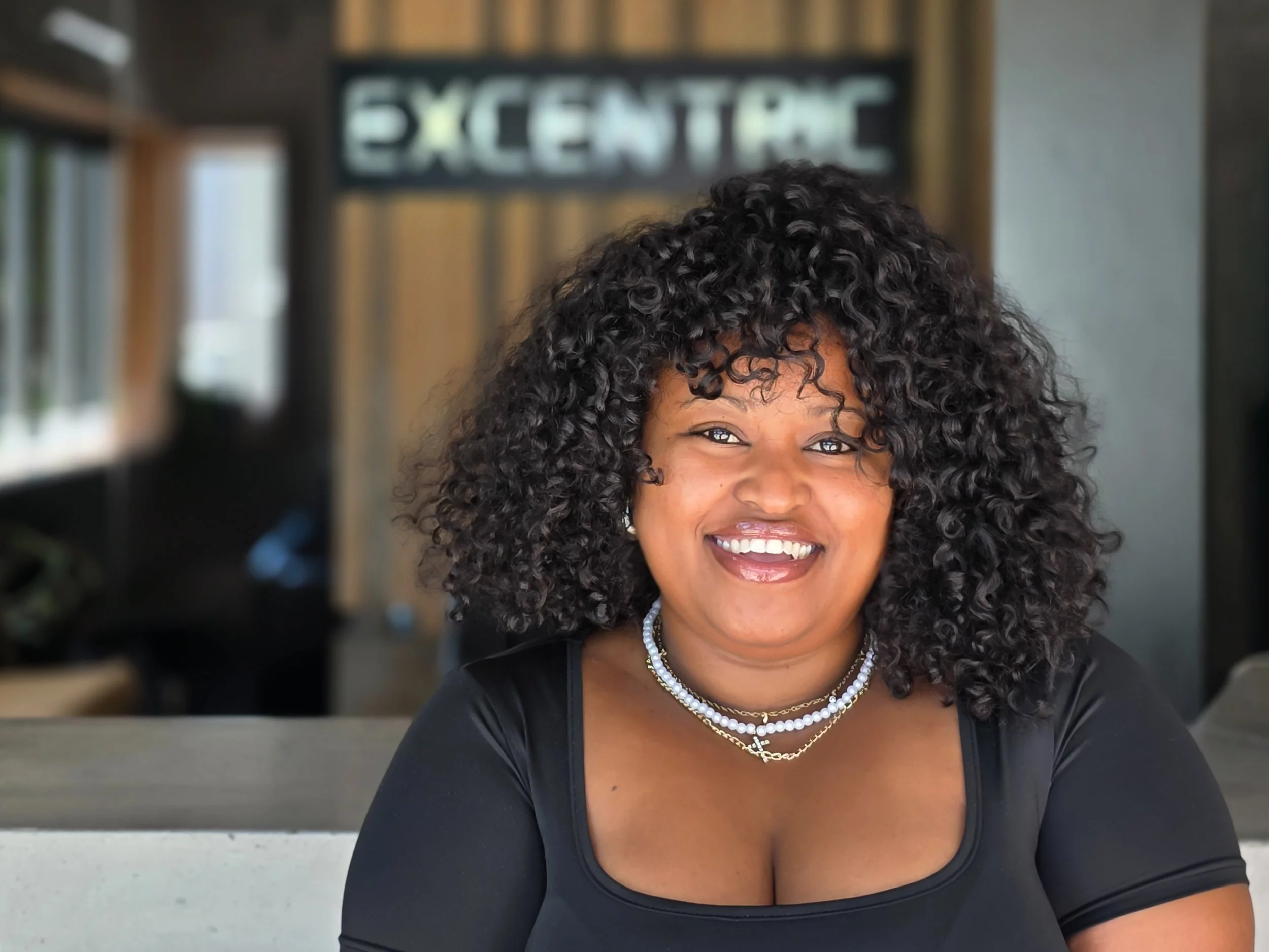 A smiling woman with curly black hair, wearing a black top and layered necklaces, seated indoors with a blurred background and a sign that reads 'EXCELLENT' behind her.