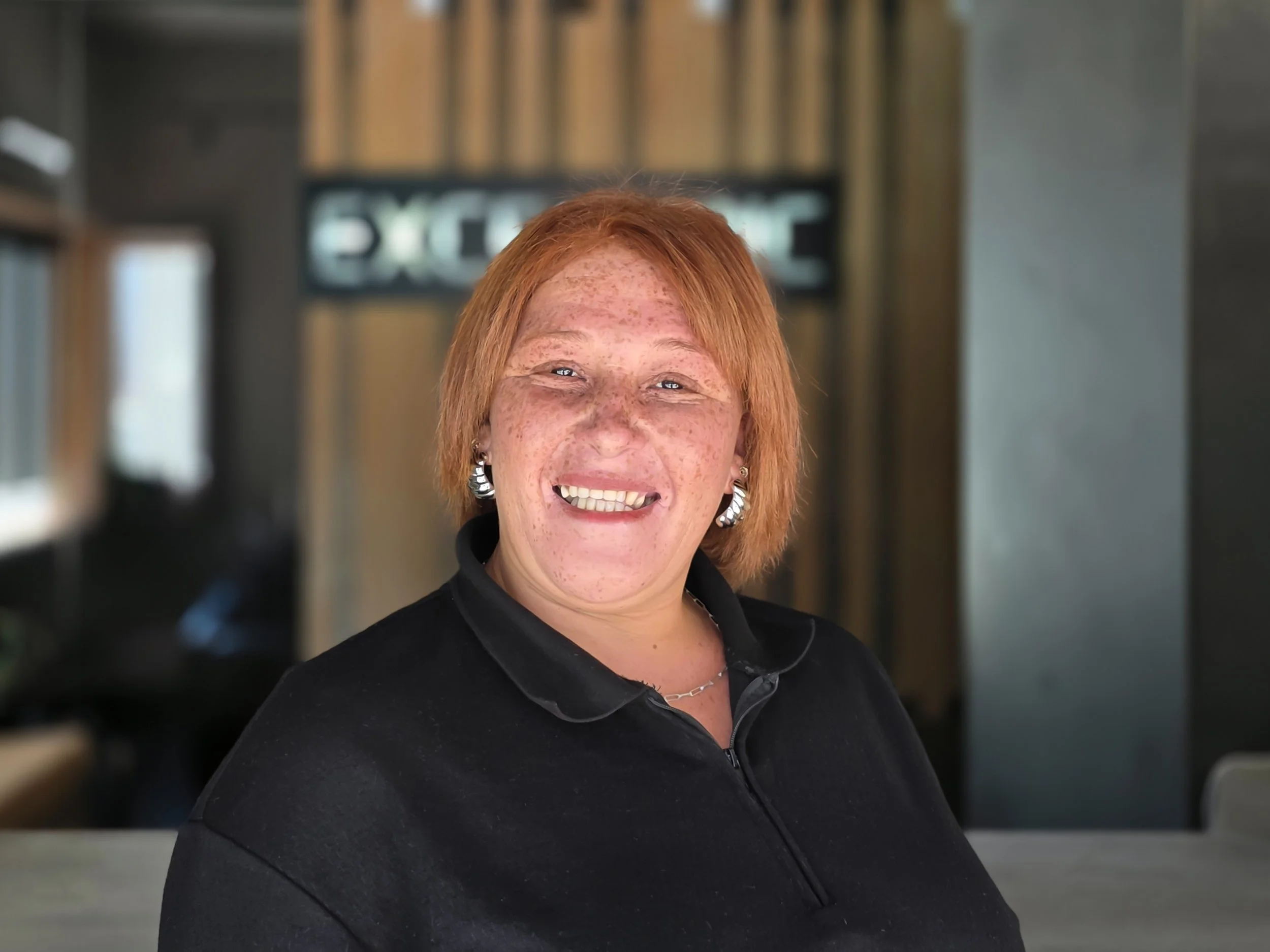 Close-up of a cheerful woman with short red hair, freckles, wearing earrings, a necklace, and a black collared shirt, in an indoor setting.