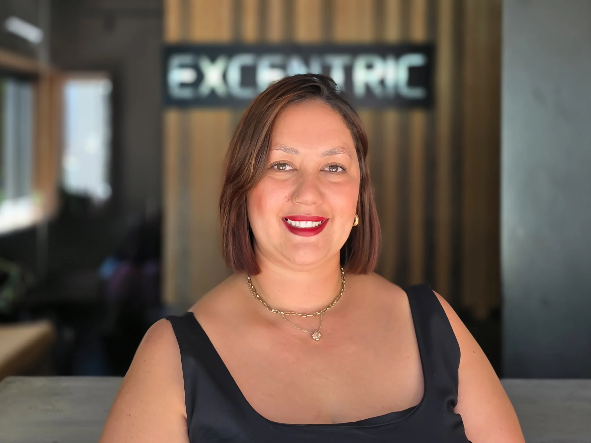 A woman with brown hair, red lipstick, wearing a black sleeveless top, gold jewelry, and a nose ring, smiling at the camera inside a modern space with wooden and dark gray walls, and a sign reading 'ELECTRIC' in the background.