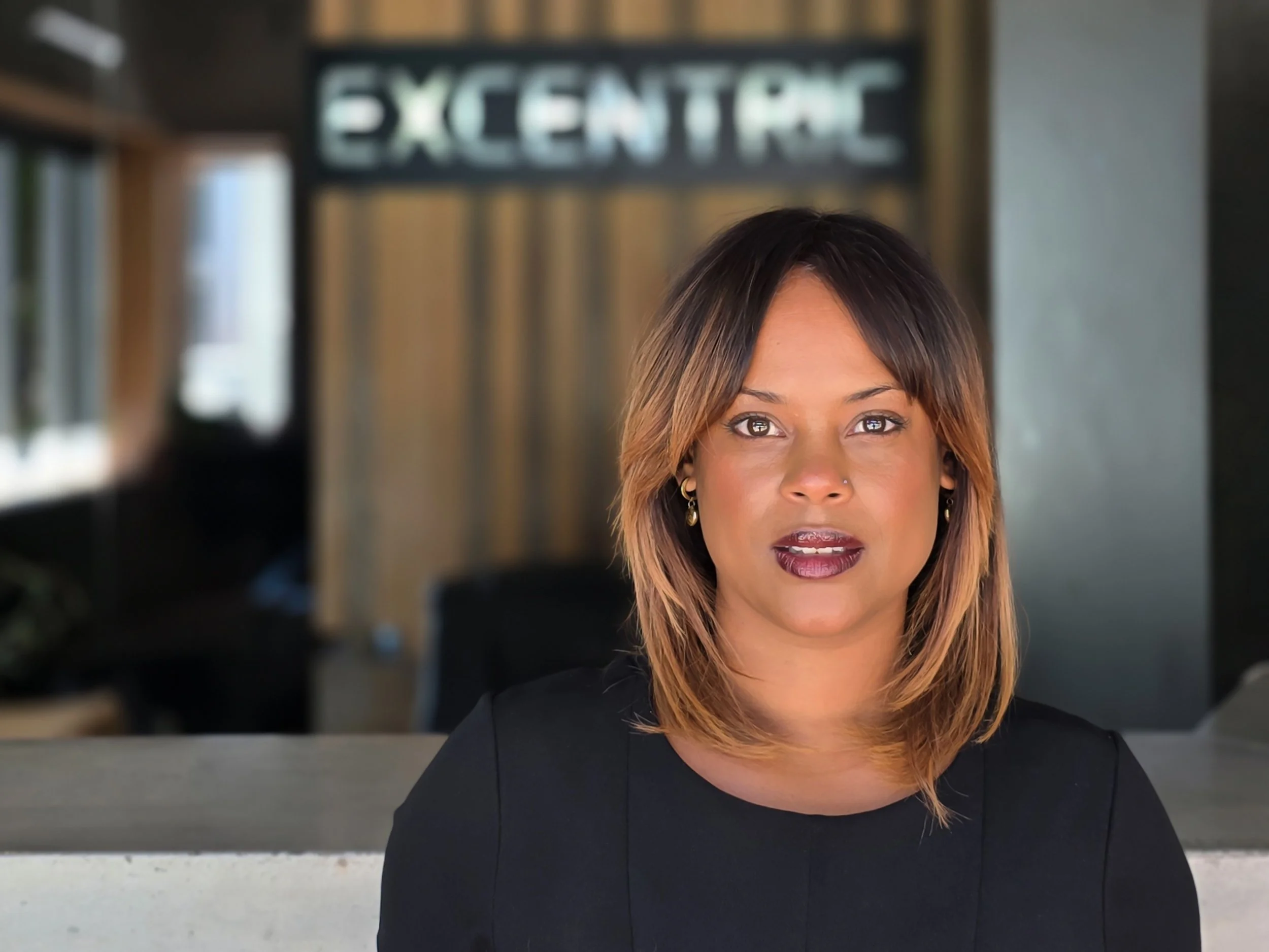 A woman with shoulder-length brown hair and dark lipstick, wearing a black shirt, sitting indoors with a sign that reads 'EXCENTRIC' in the background.