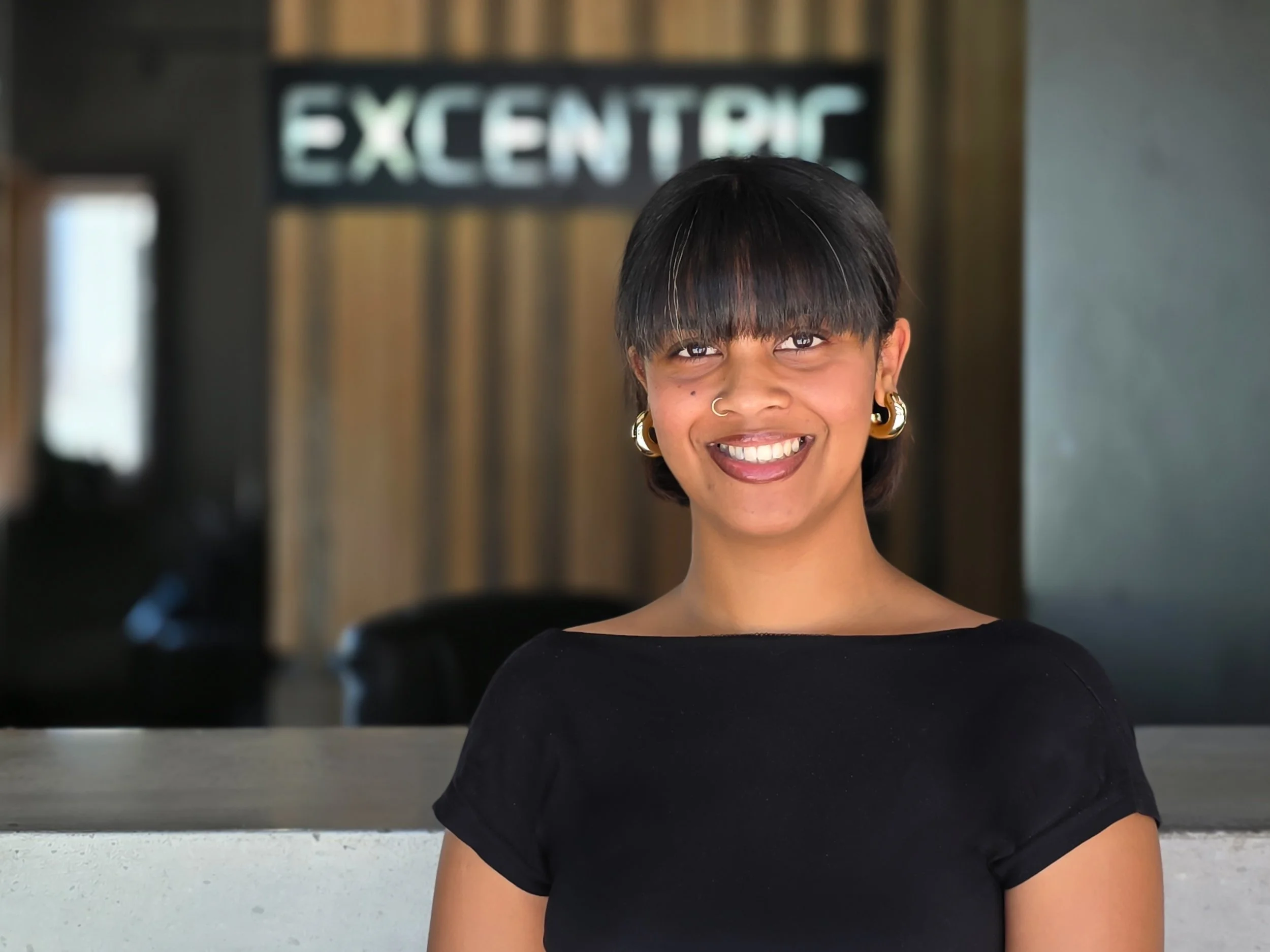 Young woman with short black hair, wearing gold earrings and a nose ring, smiling indoors in front of a blurred background with a sign that reads 'EXCENTRIC'.