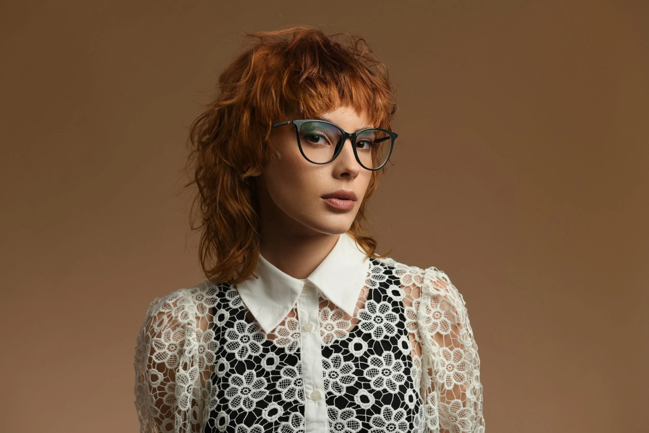 A young woman with red hair, black eyeglasses, wearing a white collared shirt under a black dress with white floral lace overlay, standing against a brown background.