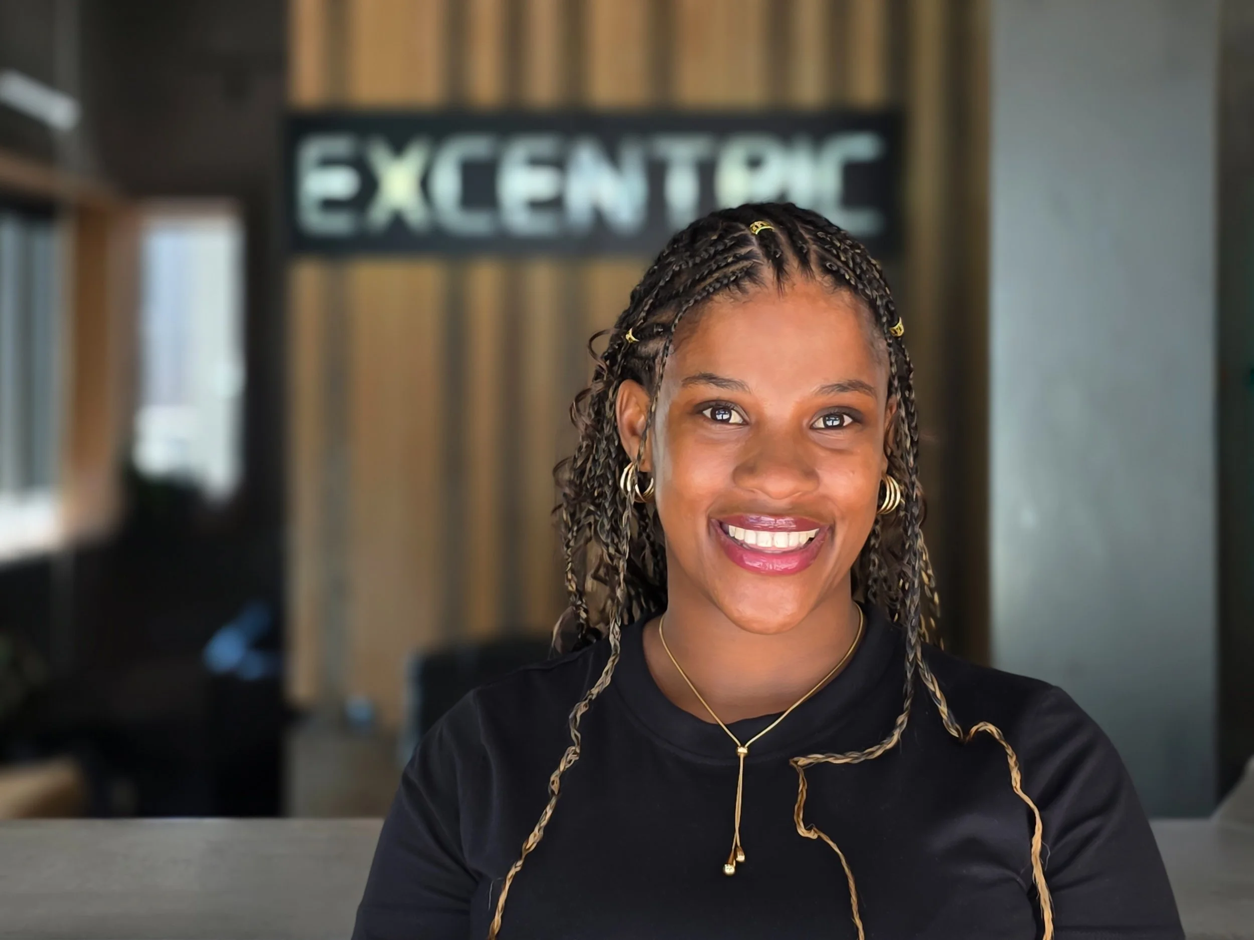 A smiling woman with braided hair and gold jewelry sitting at a table in a modern office or café with a blurred background and a sign that reads 'EXCENTRIC' behind her.