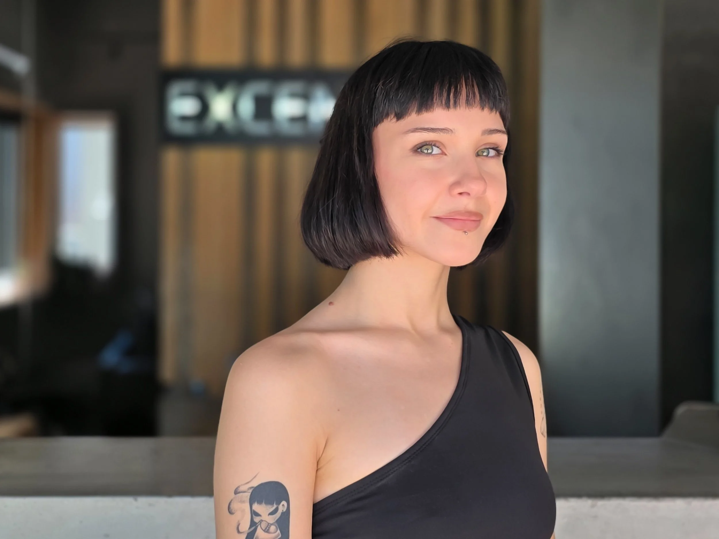 Portrait of a woman with short black hair, light-colored eyes, a small piercing below her lower lip, tattoos on her upper arms, wearing a black sleeveless top, standing indoors with a blurred background.