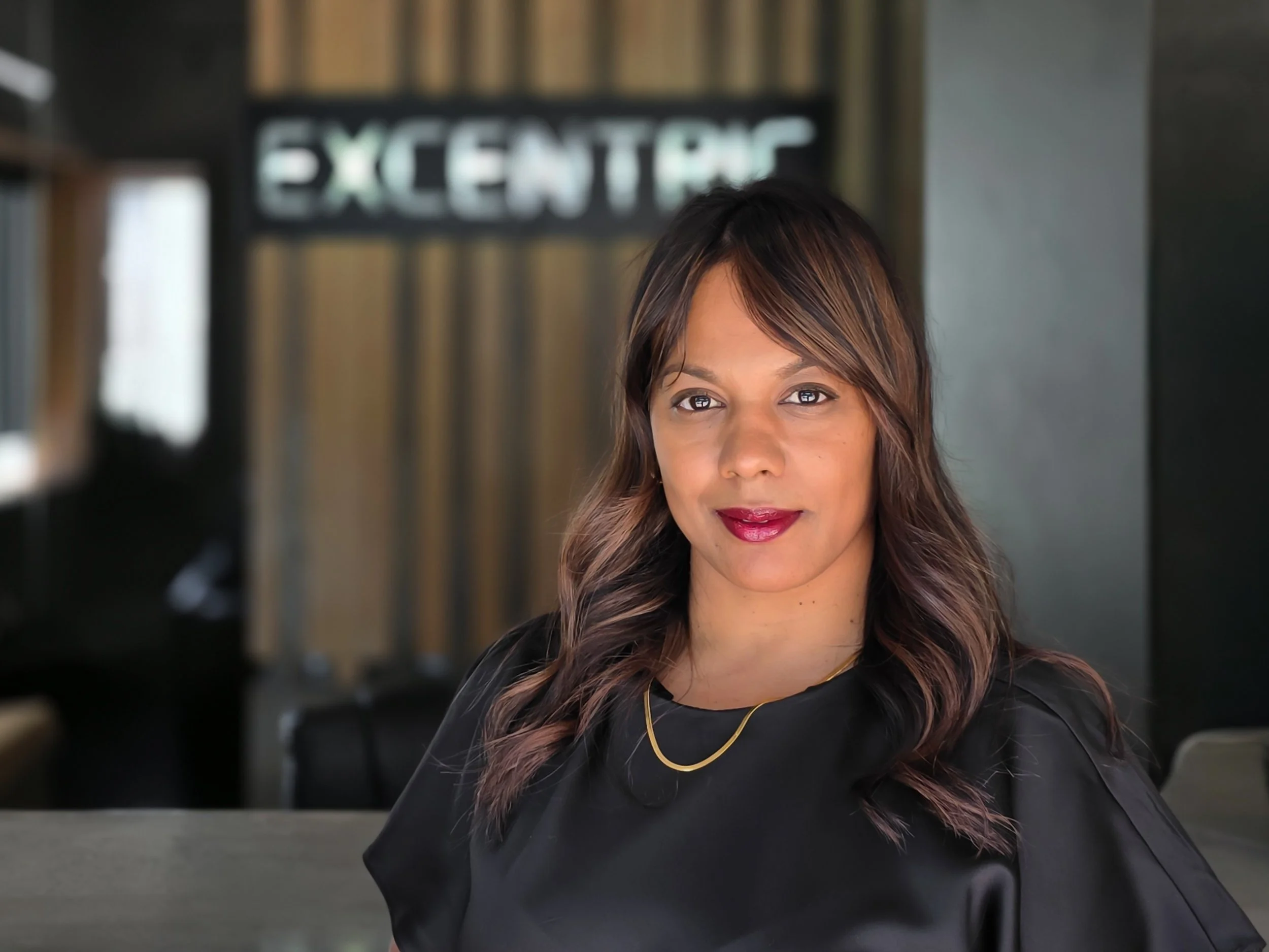A woman with shoulder-length brown hair and a black top, smiling in an indoor setting, with a blurred background and a sign that reads 'EXCENTRIC'.