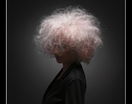 Side profile of a person with curly, disheveled, white hair against a dark background.
