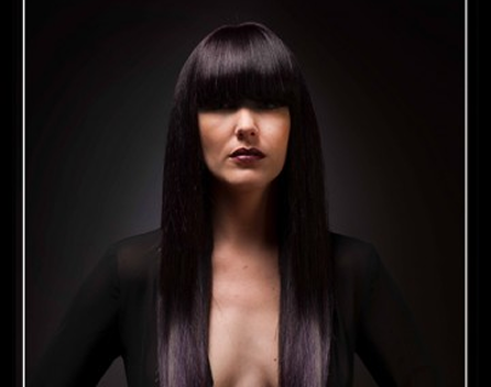 A woman with long, straight black hair and blunt bangs, wearing dark makeup and a black top, against a dark background.