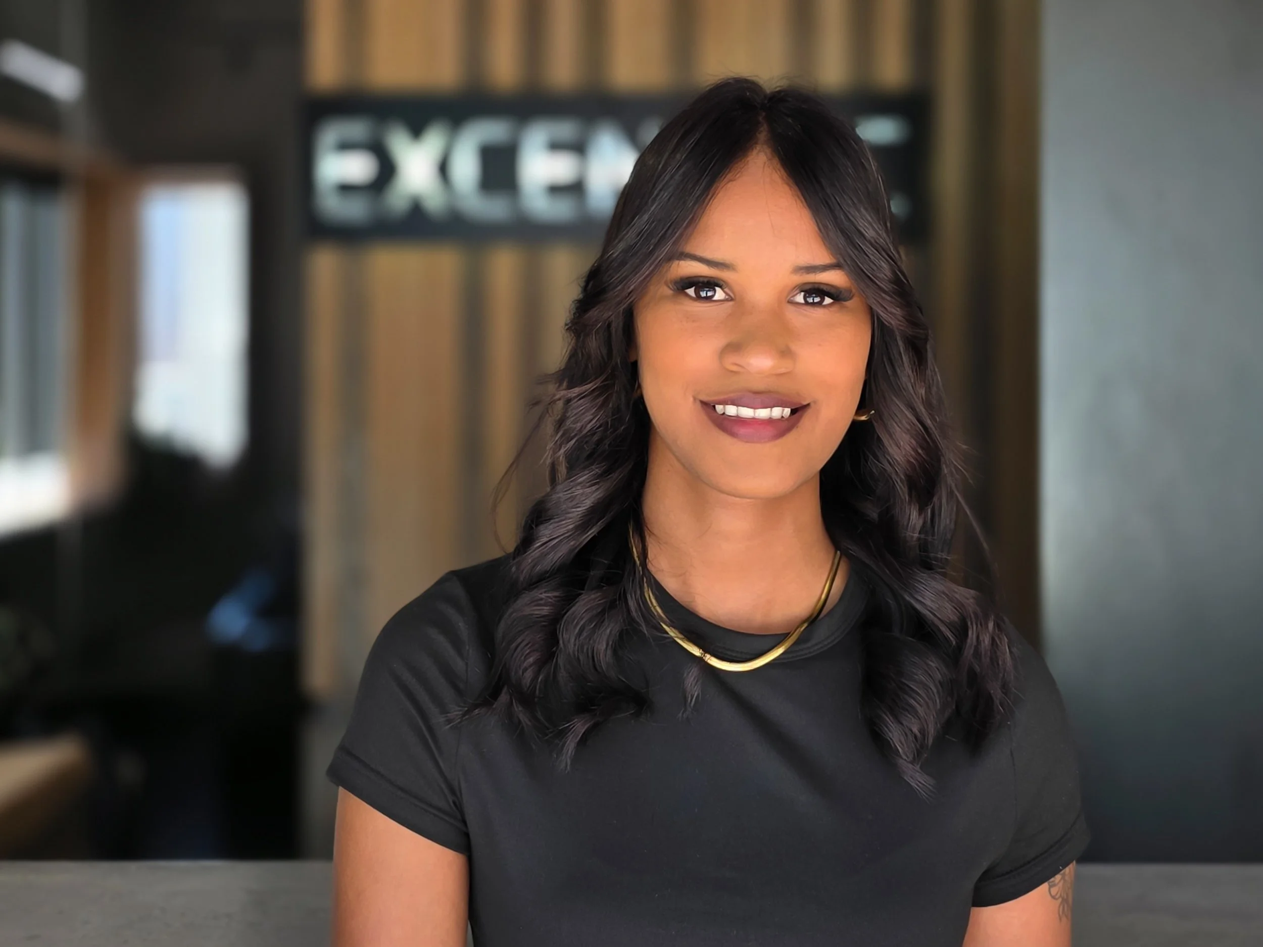 A woman with long, dark, wavy hair smiling at the camera, wearing a black shirt and gold jewelry, in an indoor setting with a blurred background featuring a sign that reads 'EXCELLENCE'.