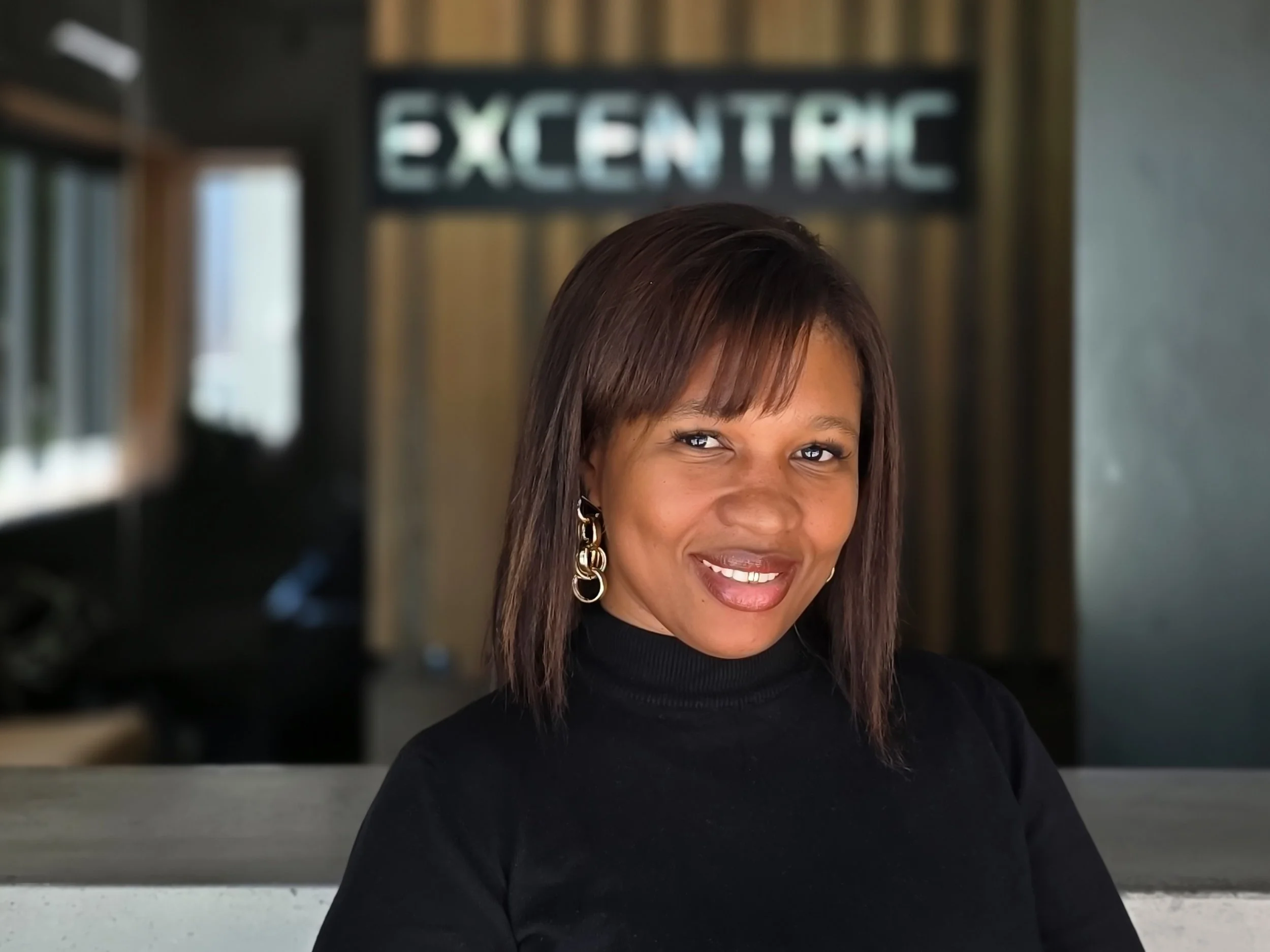 Close-up of a woman with shoulder-length brown hair, wearing gold hoop earrings and a black turtleneck, smiling at the camera, with a background sign that reads 'EXCENTRIC'.