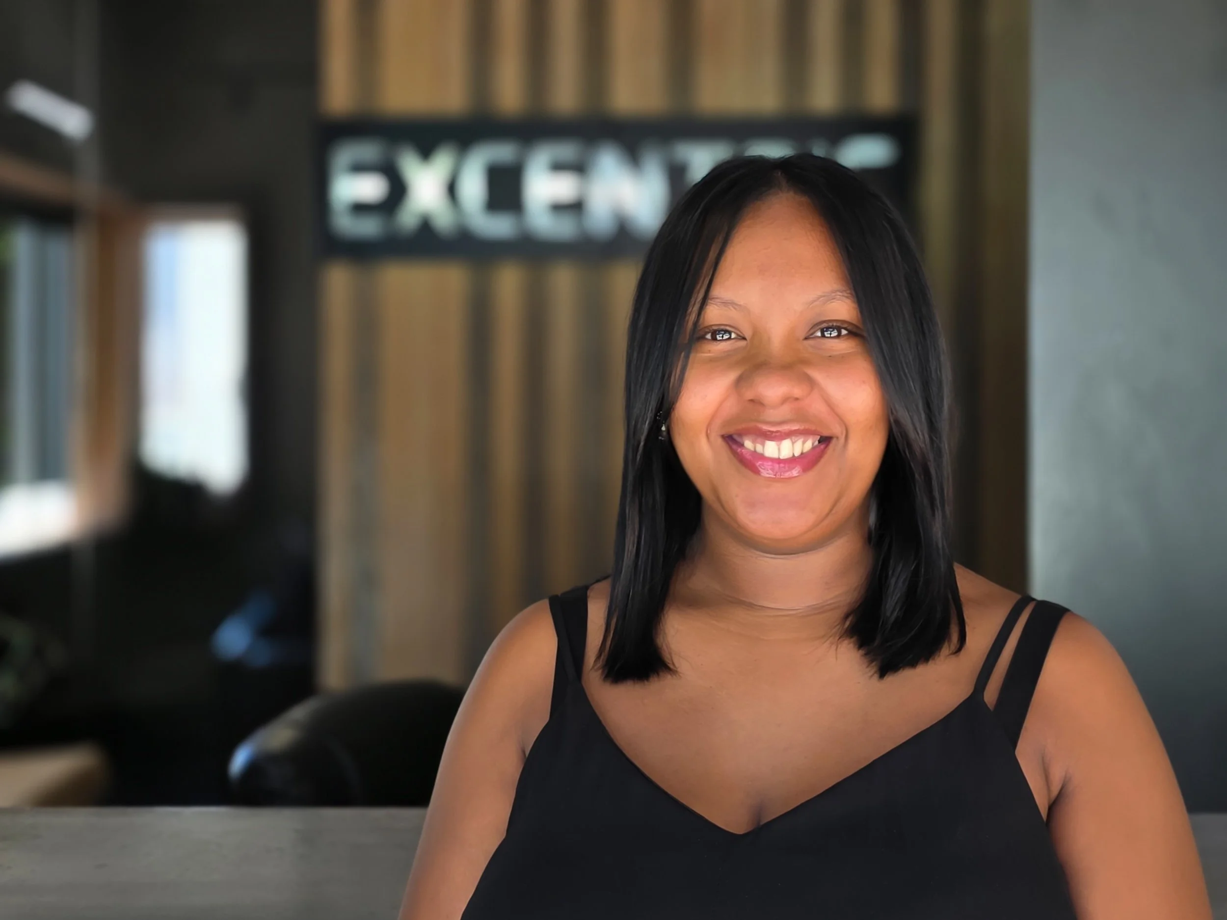 A smiling woman with medium-length black hair, wearing a black sleeveless top, standing indoors with a blurred background featuring a sign that reads 'EXCELLENT' and some wooden and metal decor.