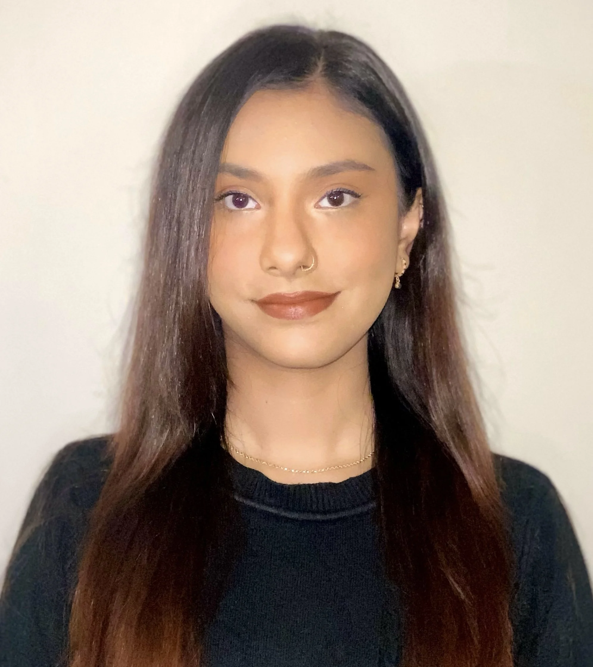 A young woman with long dark brown hair, wearing a black top, gold jewelry, and makeup, smiling at the camera against a plain light-colored background.