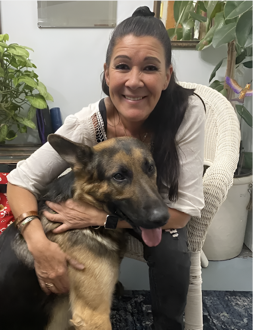 A woman smiling and hugging a German Shepherd dog indoors surrounded by plants and home decor.
