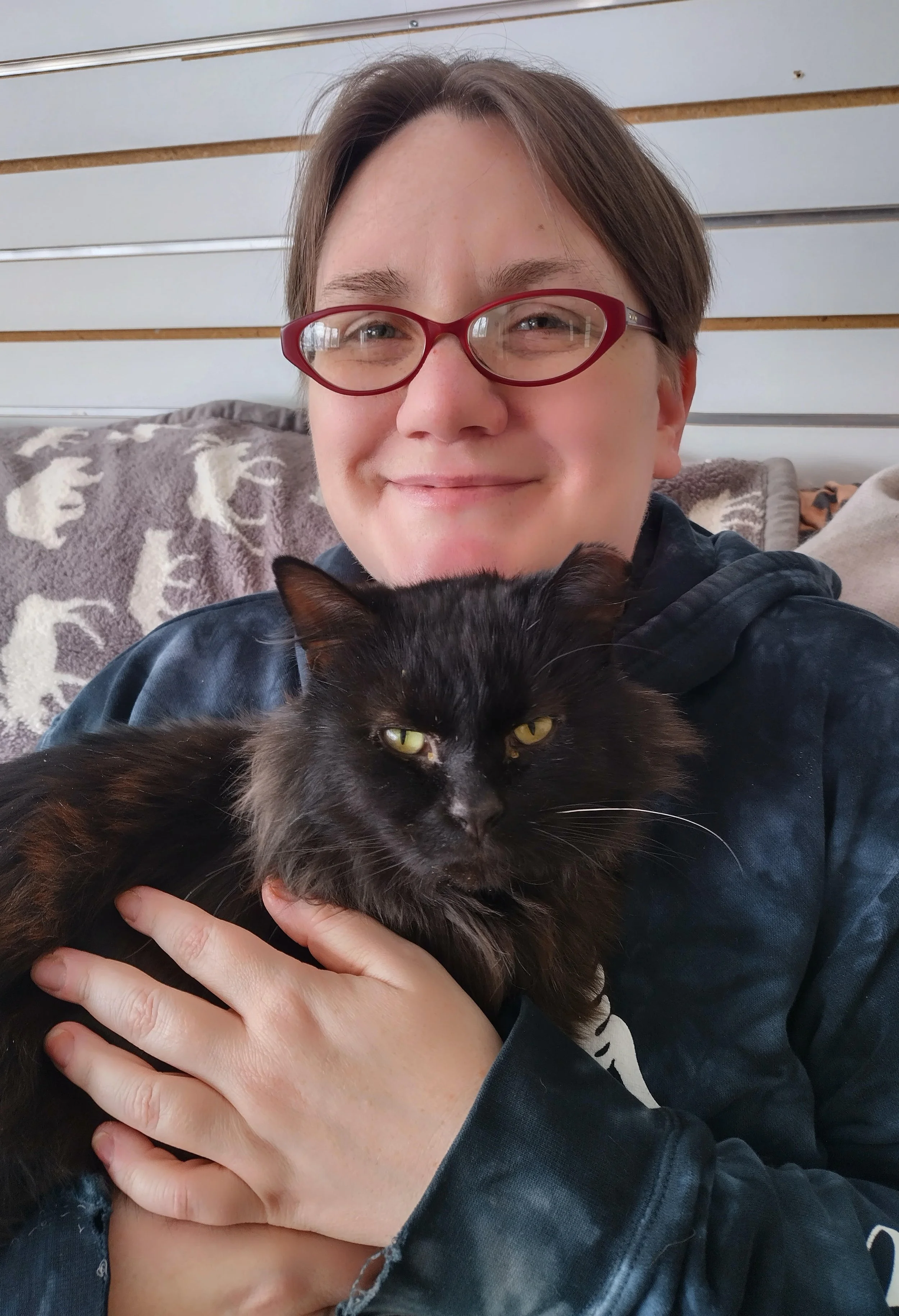 A person with short brown hair, wearing red glasses, smiling while holding a long-haired black cat with yellow eyes.