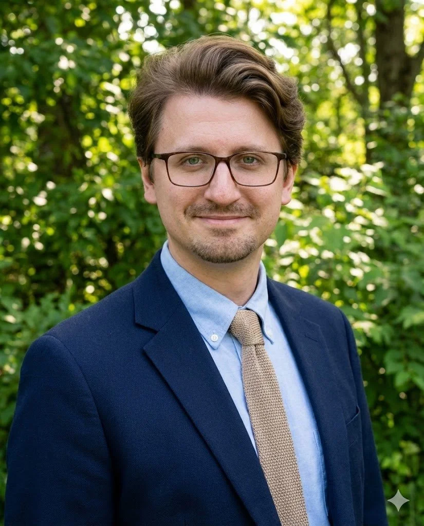 Portrait of a man with glasses, wearing a navy blazer, light blue dress shirt, and beige tie, standing outdoors with green foliage in the background.