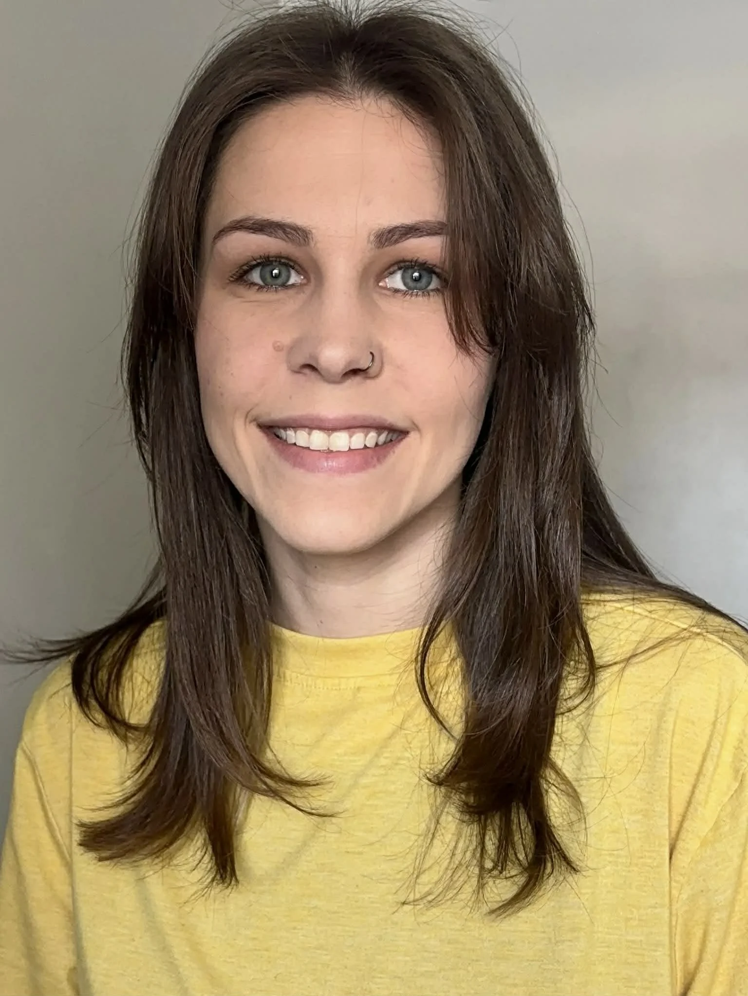 A young woman with blue eyes, light skin, and brown hair, wearing a yellow shirt, smiling at the camera against a plain background.