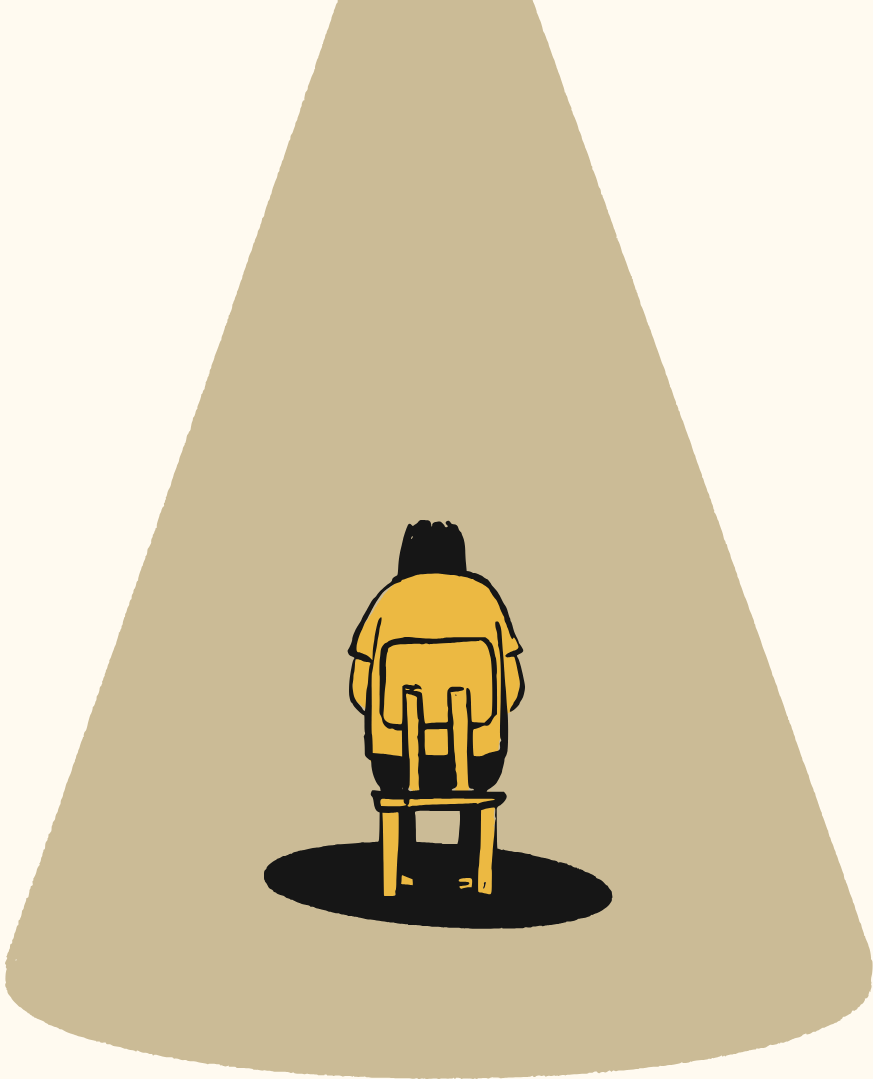 A person sitting alone under a spotlight in a dark, empty space, facing away from the viewer, wearing a yellow coat and a backpack.