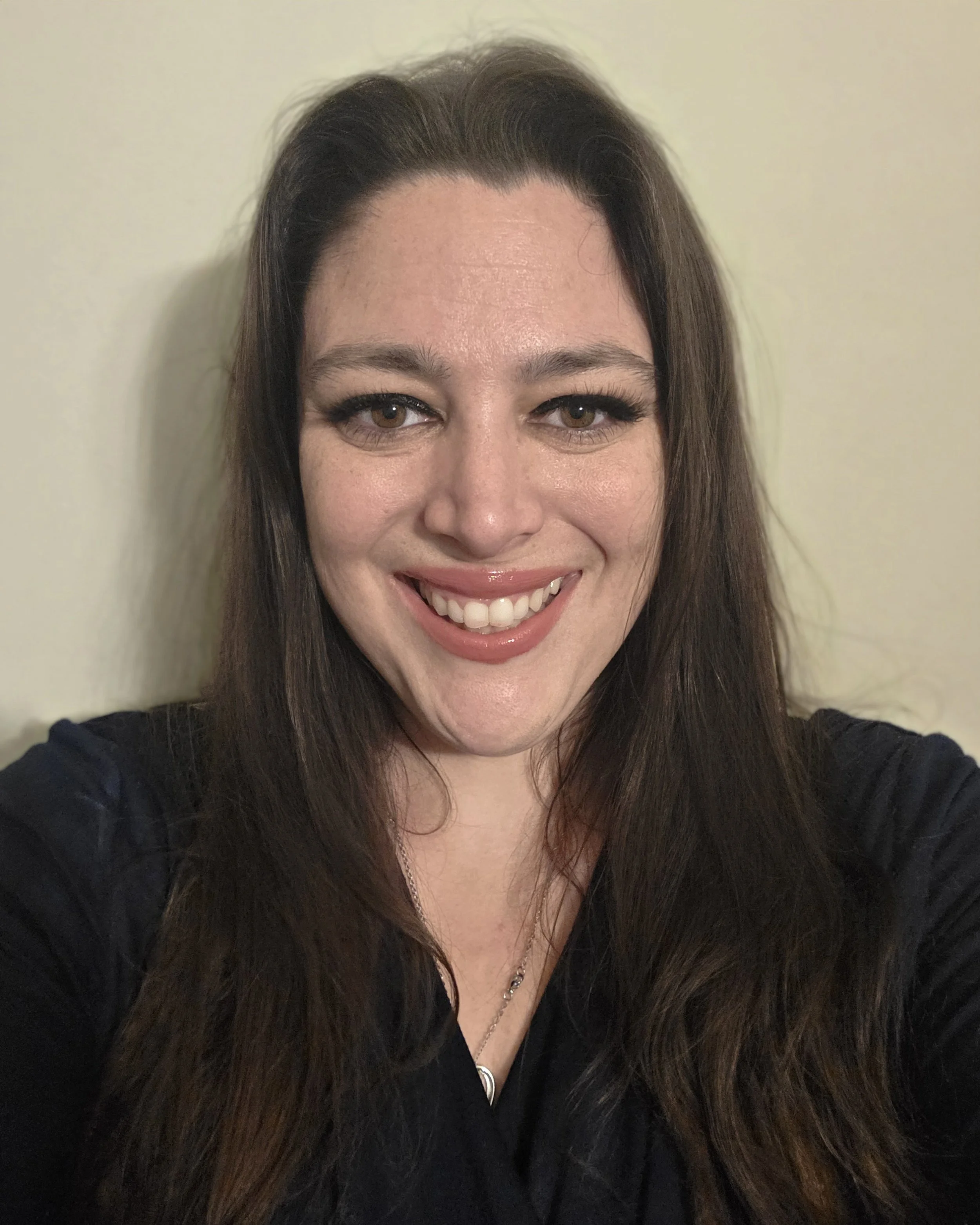 A woman with long brown hair smiling and wearing a black top, standing in front of a plain, light-colored wall.