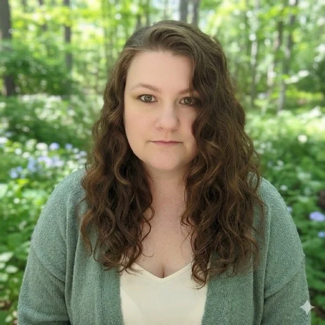Portrait of a woman with long, curly brown hair, wearing a white top and a green cardigan, standing outdoors in a wooded area with green foliage in the background.